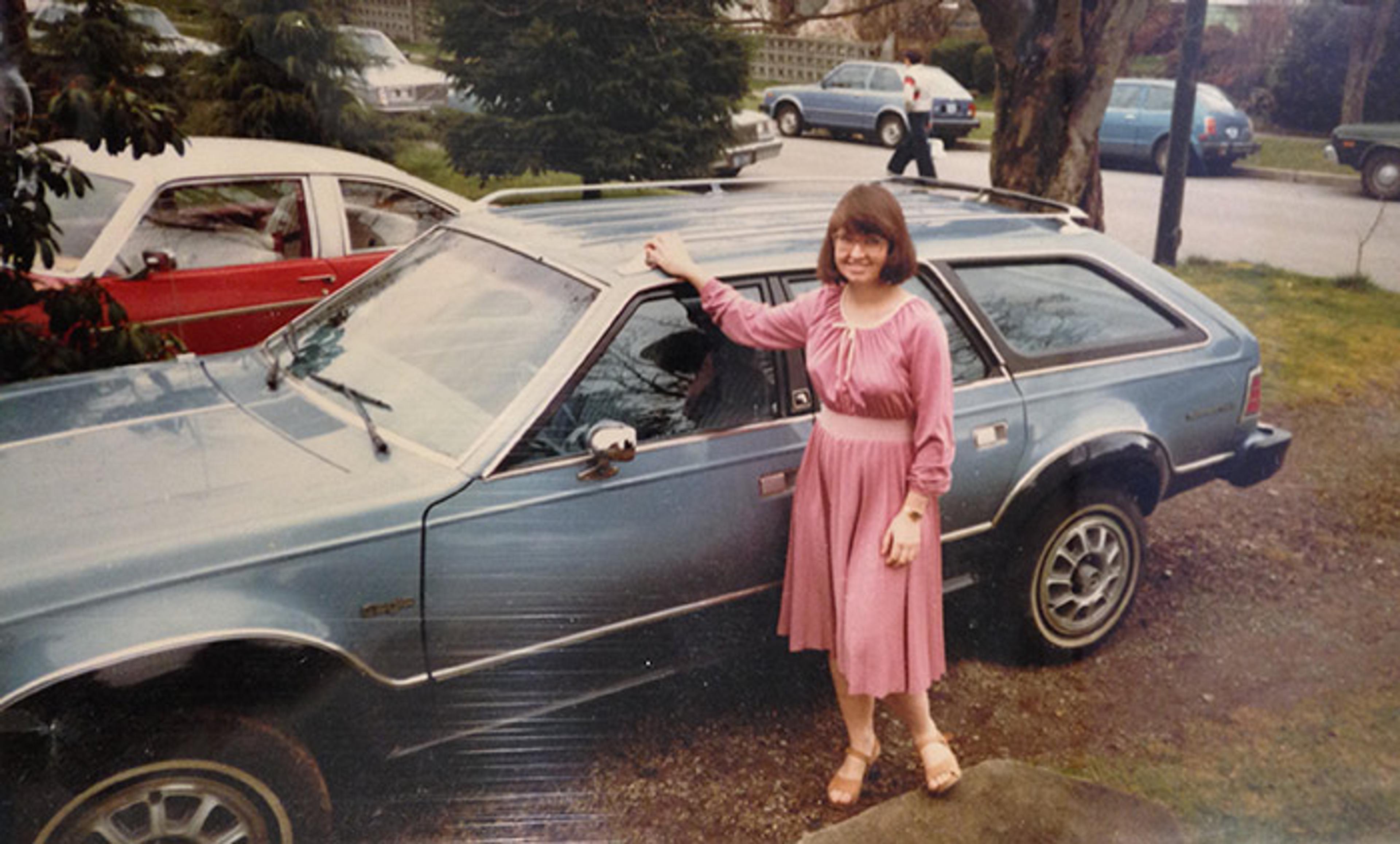 Vintage photo of a woman in a pink dress standing next to a blue car, with other cars and trees in the background.