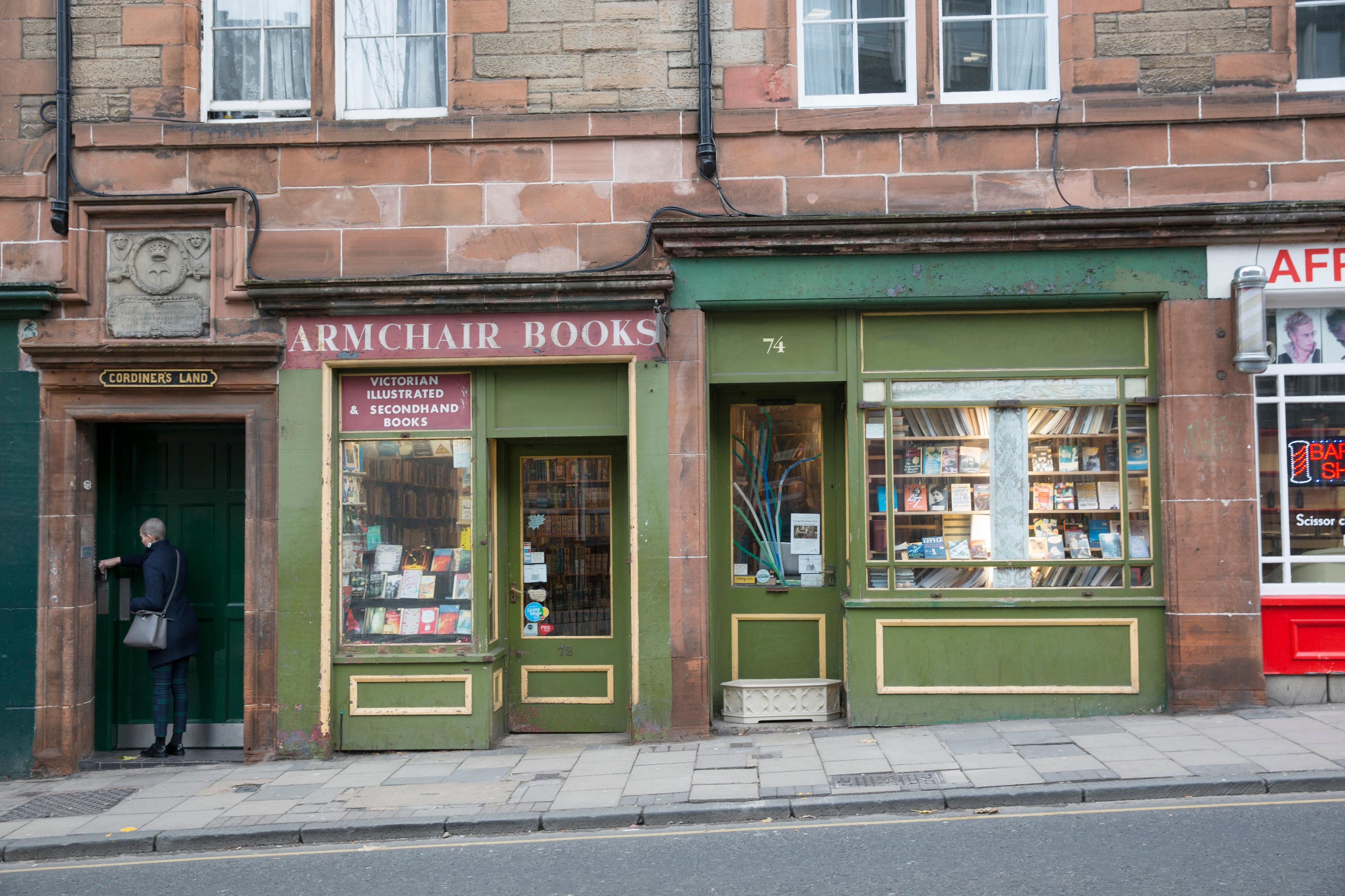Photo of a quaint bookshop with a green facade and display window filled with books, a person entering via a side door.