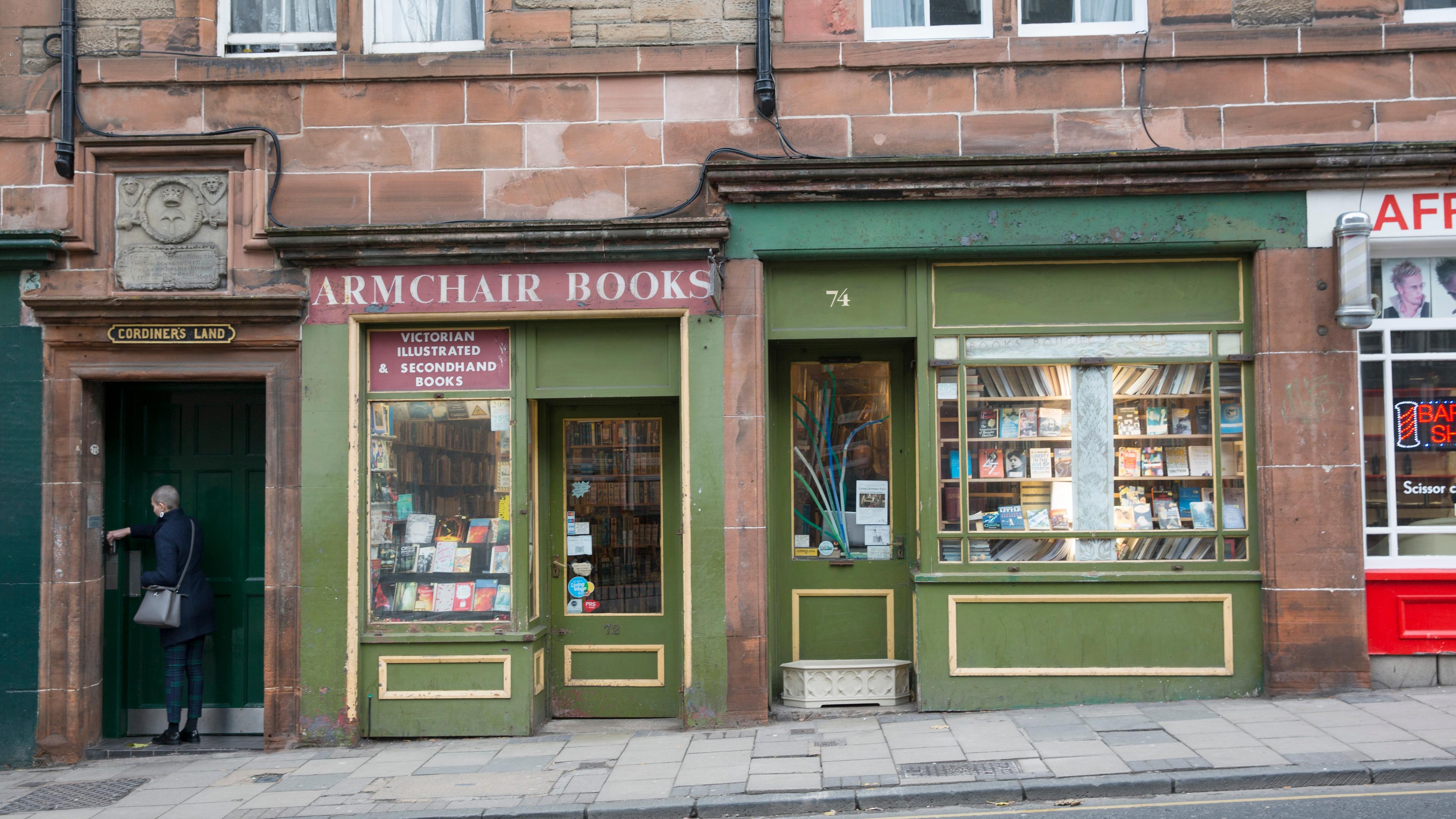 Photo of a quaint bookshop with a green facade and display window filled with books, a person entering via a side door.