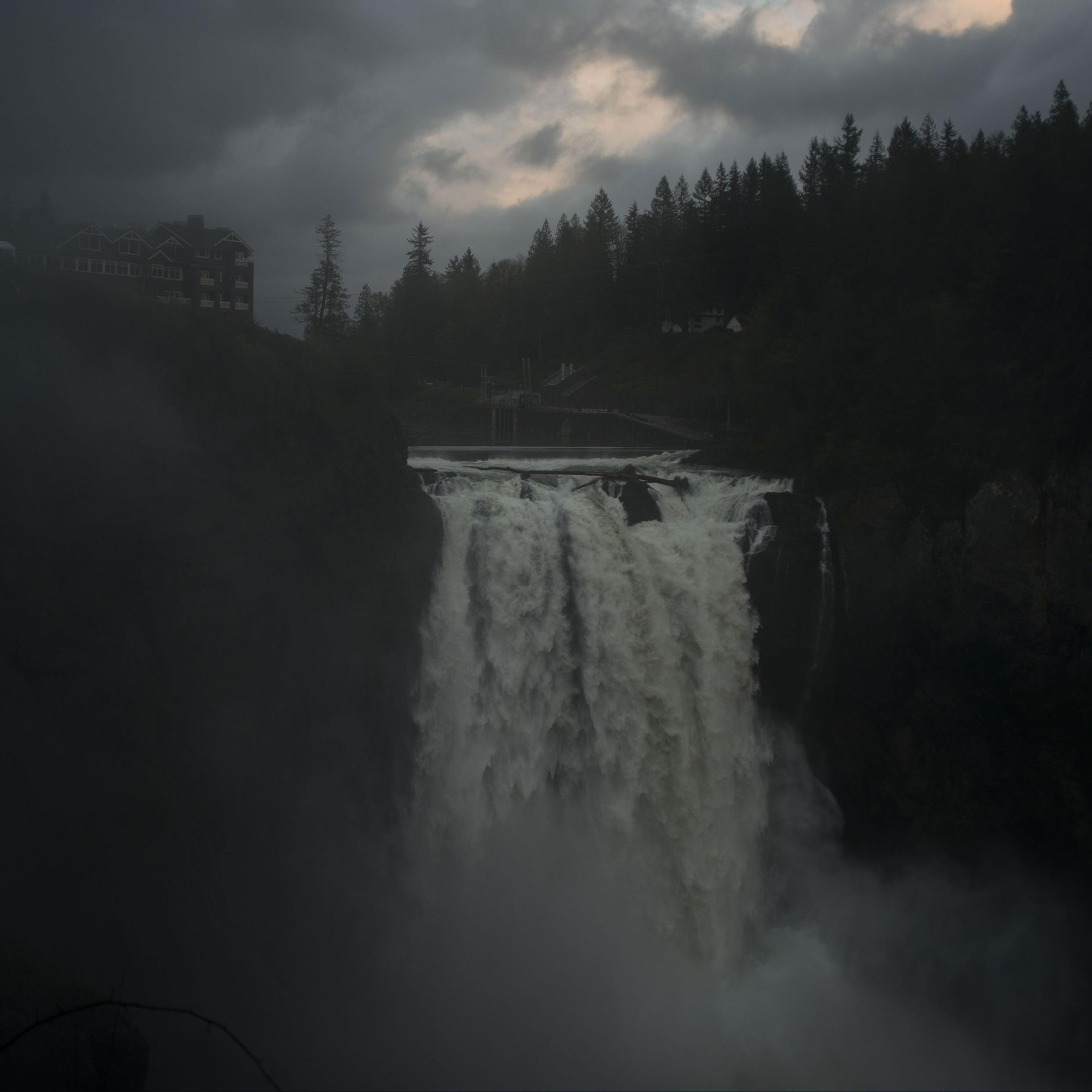 A waterfall under a stormy sky with mist and a building on the left cliff surrounded by trees in the background.