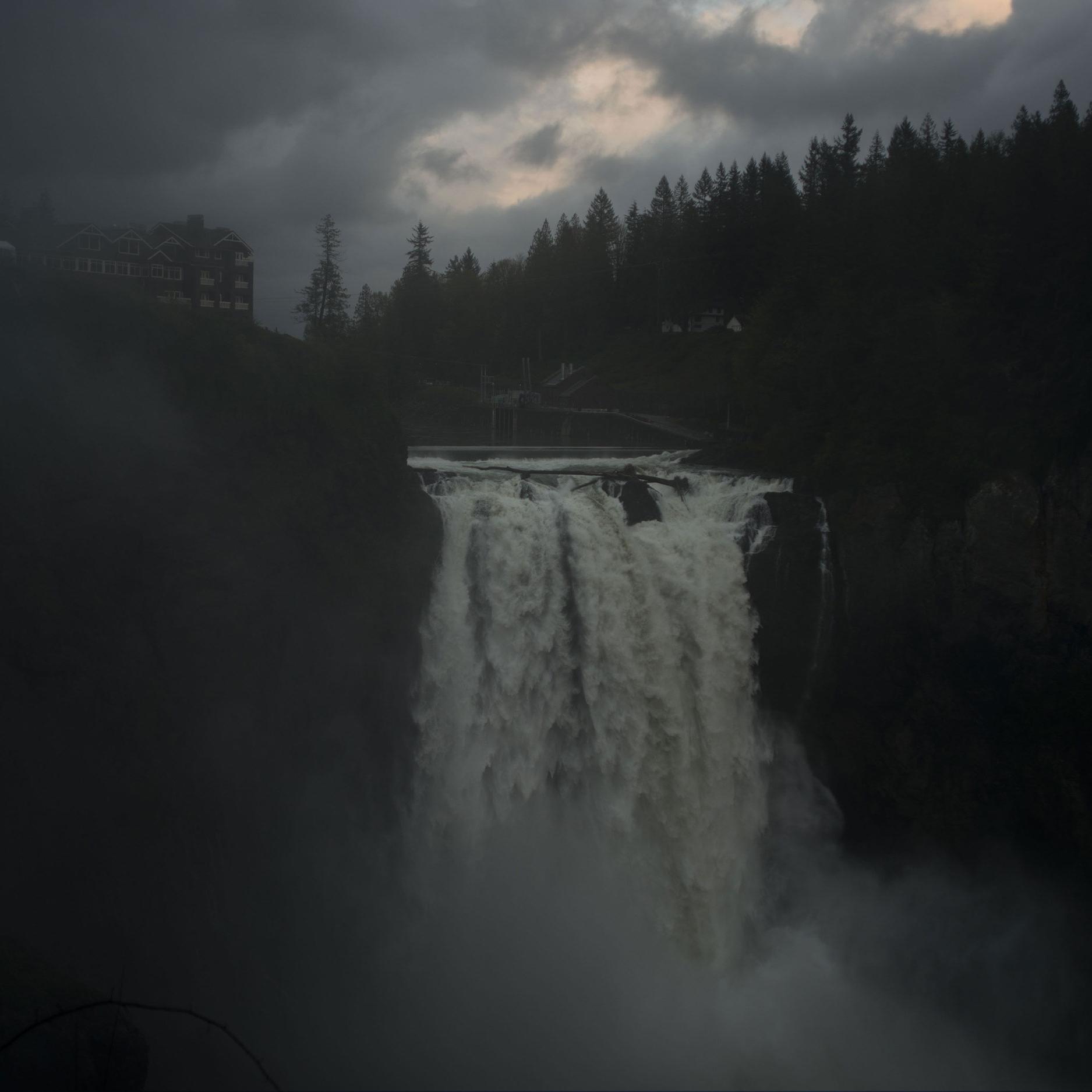 A waterfall under a stormy sky with mist and a building on the left cliff surrounded by trees in the background.
