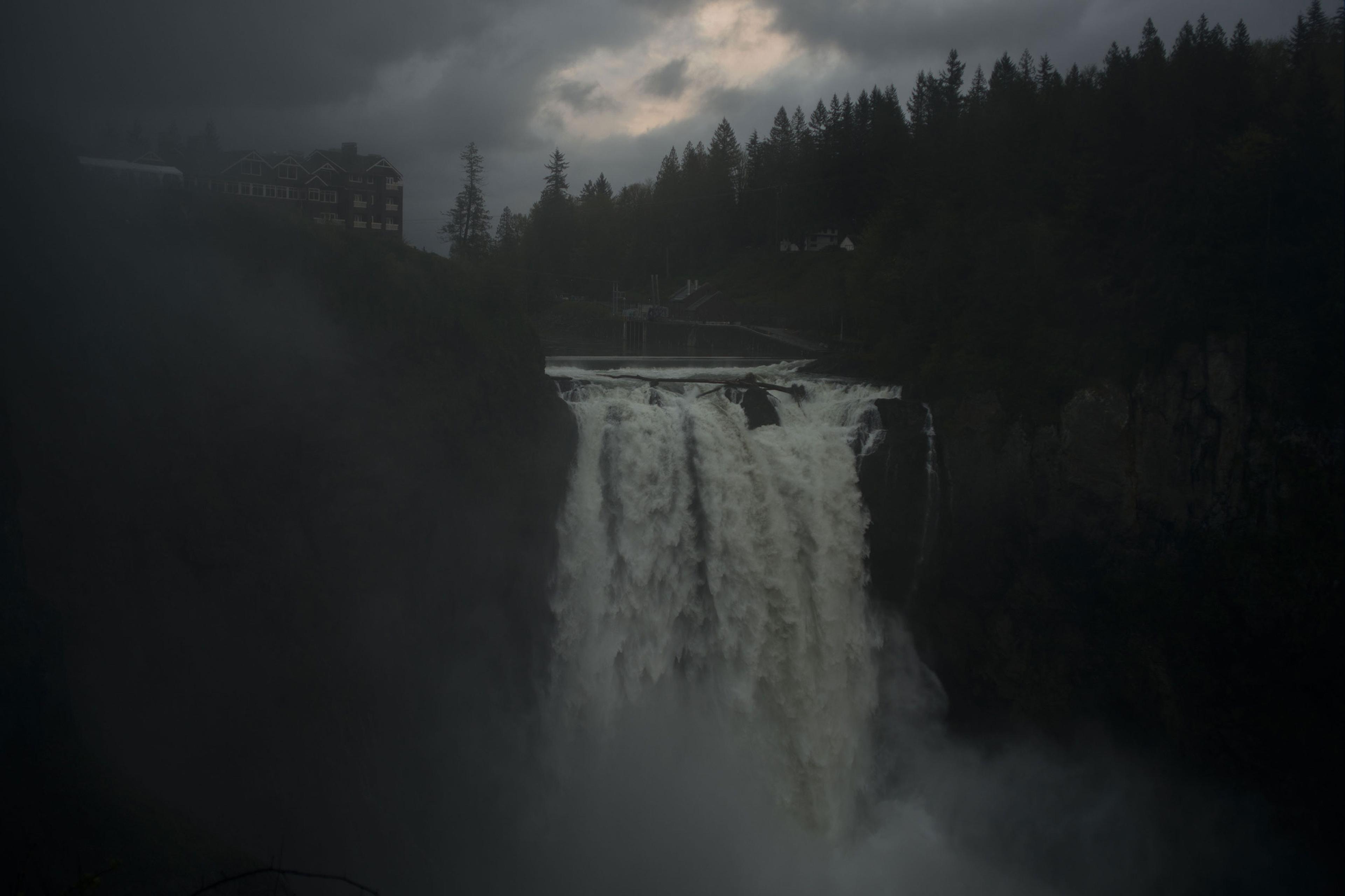 A waterfall under a stormy sky with mist and a building on the left cliff surrounded by trees in the background.
