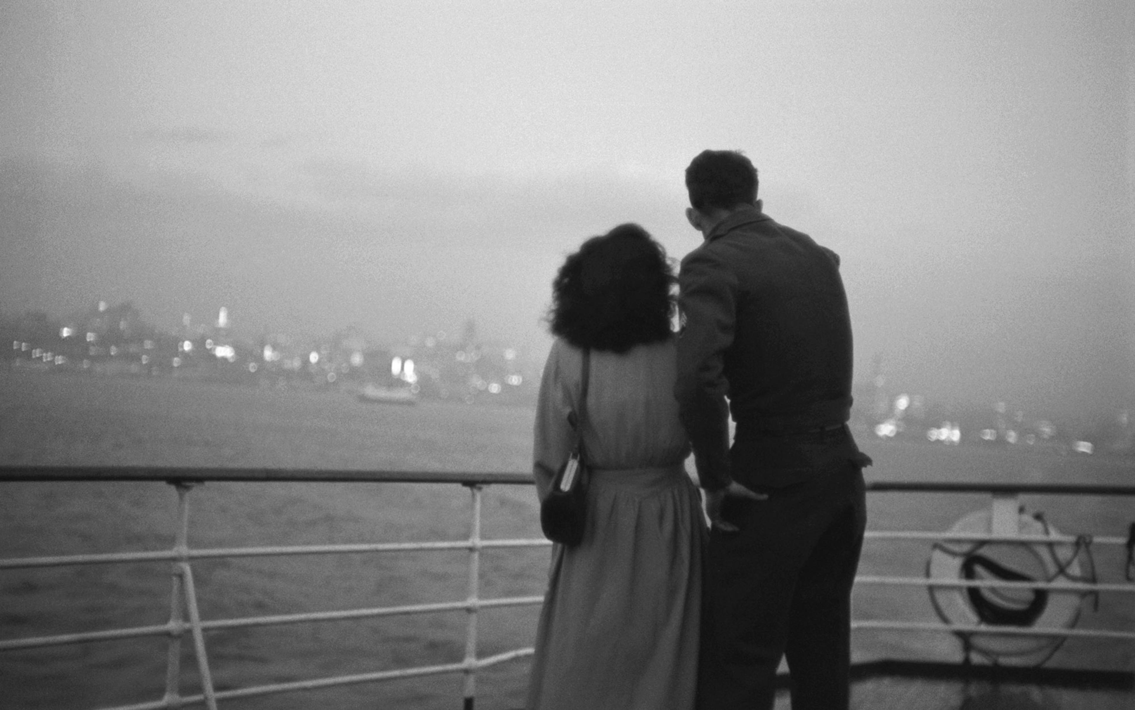 Black-and-white photo of a man and a woman, seen from behind, on the deck of a boat, looking out to shore