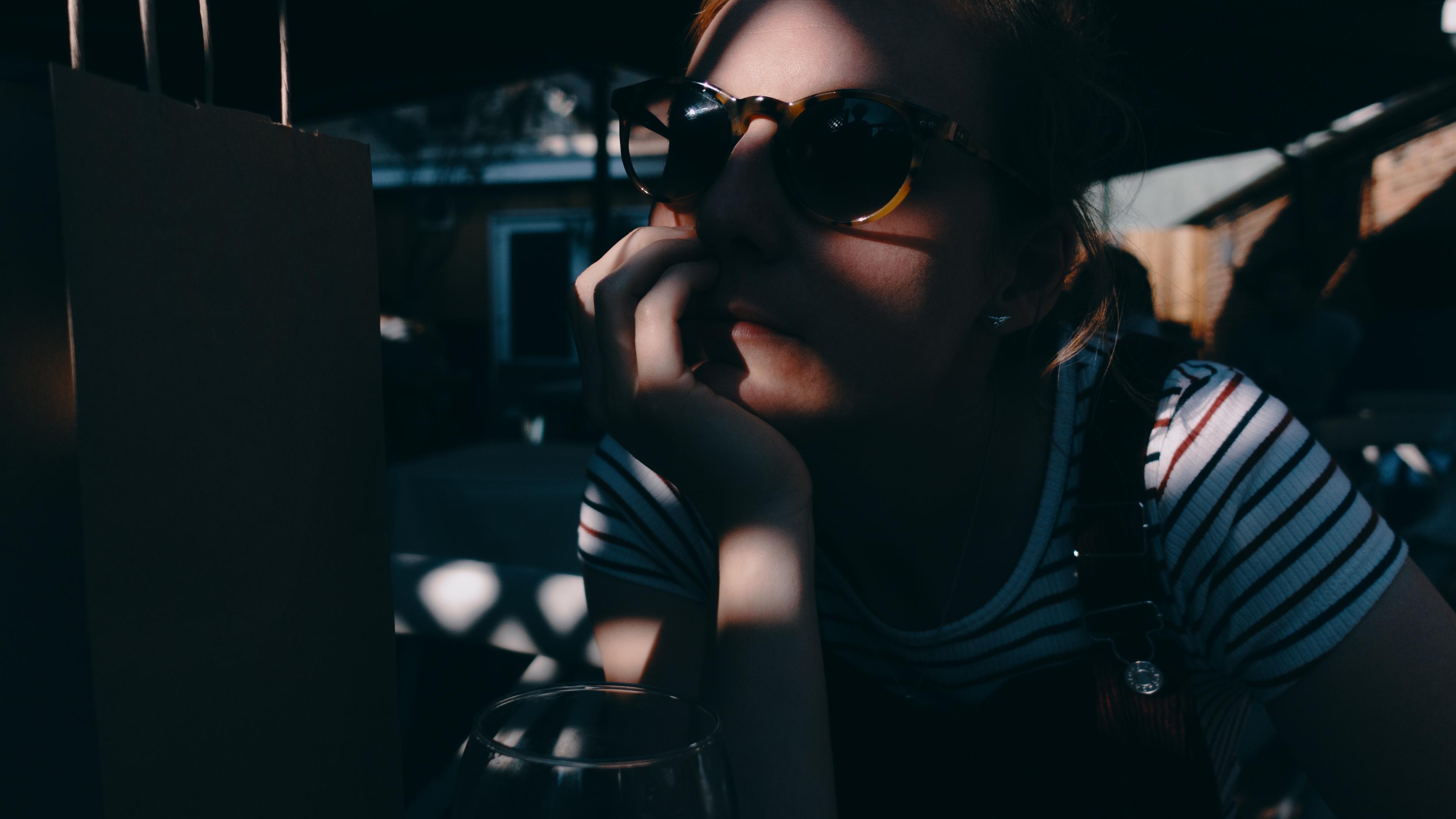 Photo of a person wearing sunglasses sitting in dappled sunlight with a thoughtful expression. Shadows cast on the face.