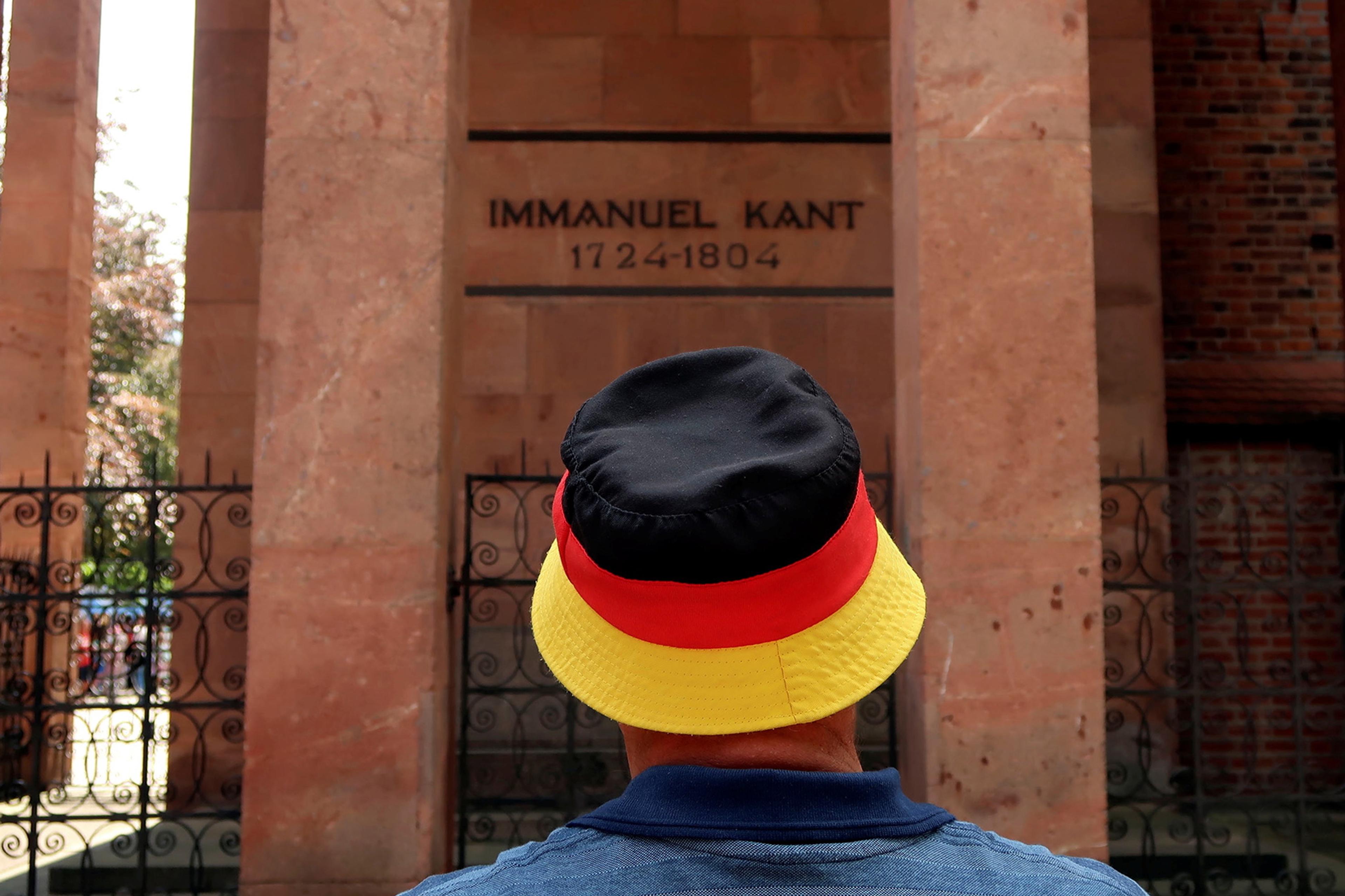 Photo of a person wearing a hat with German flag colours stands before Immanuel Kant’s memorial inscribed with dates 1724-1804.