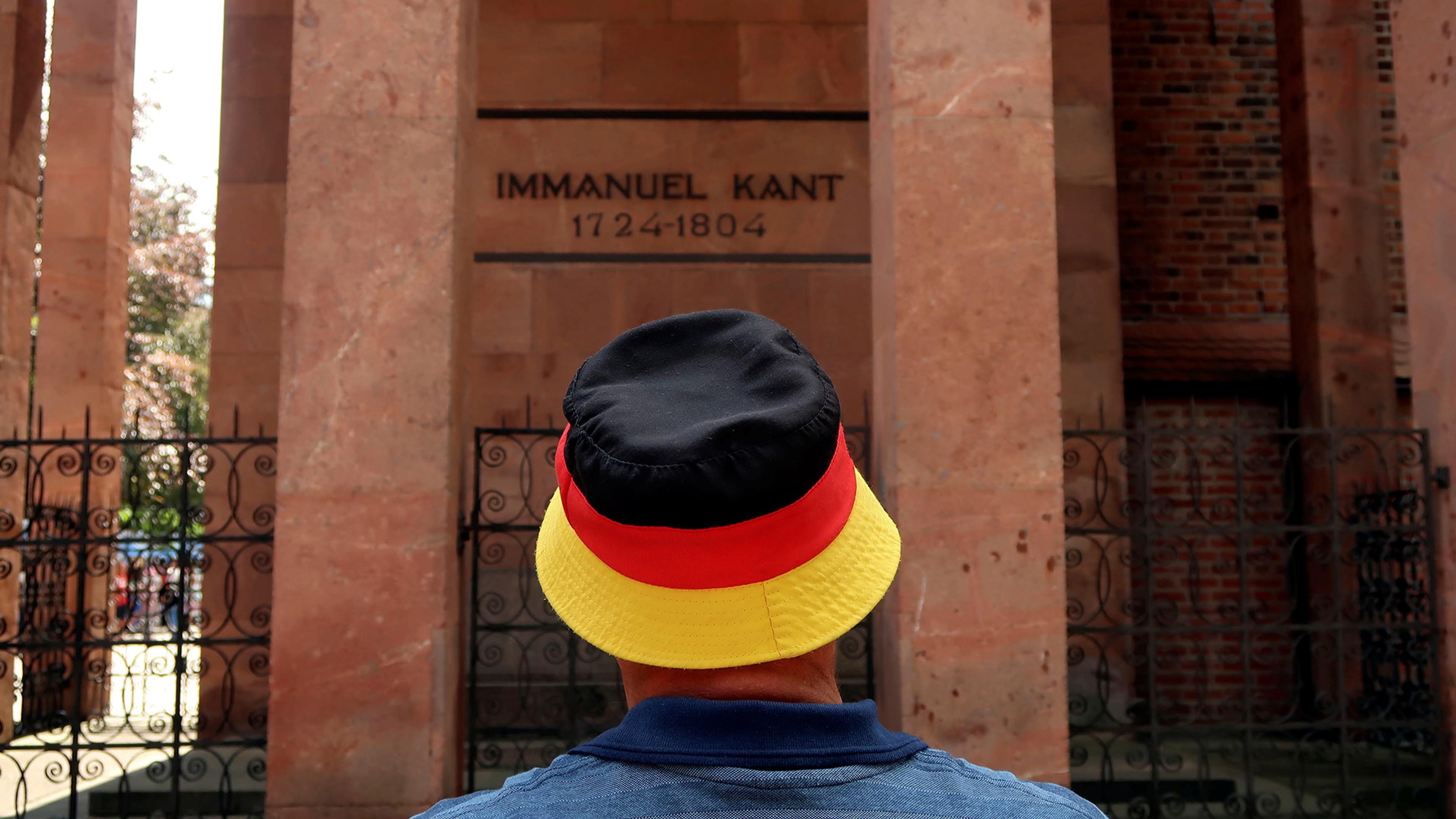 Photo of a person wearing a hat with German flag colours stands before Immanuel Kant’s memorial inscribed with dates 1724-1804.