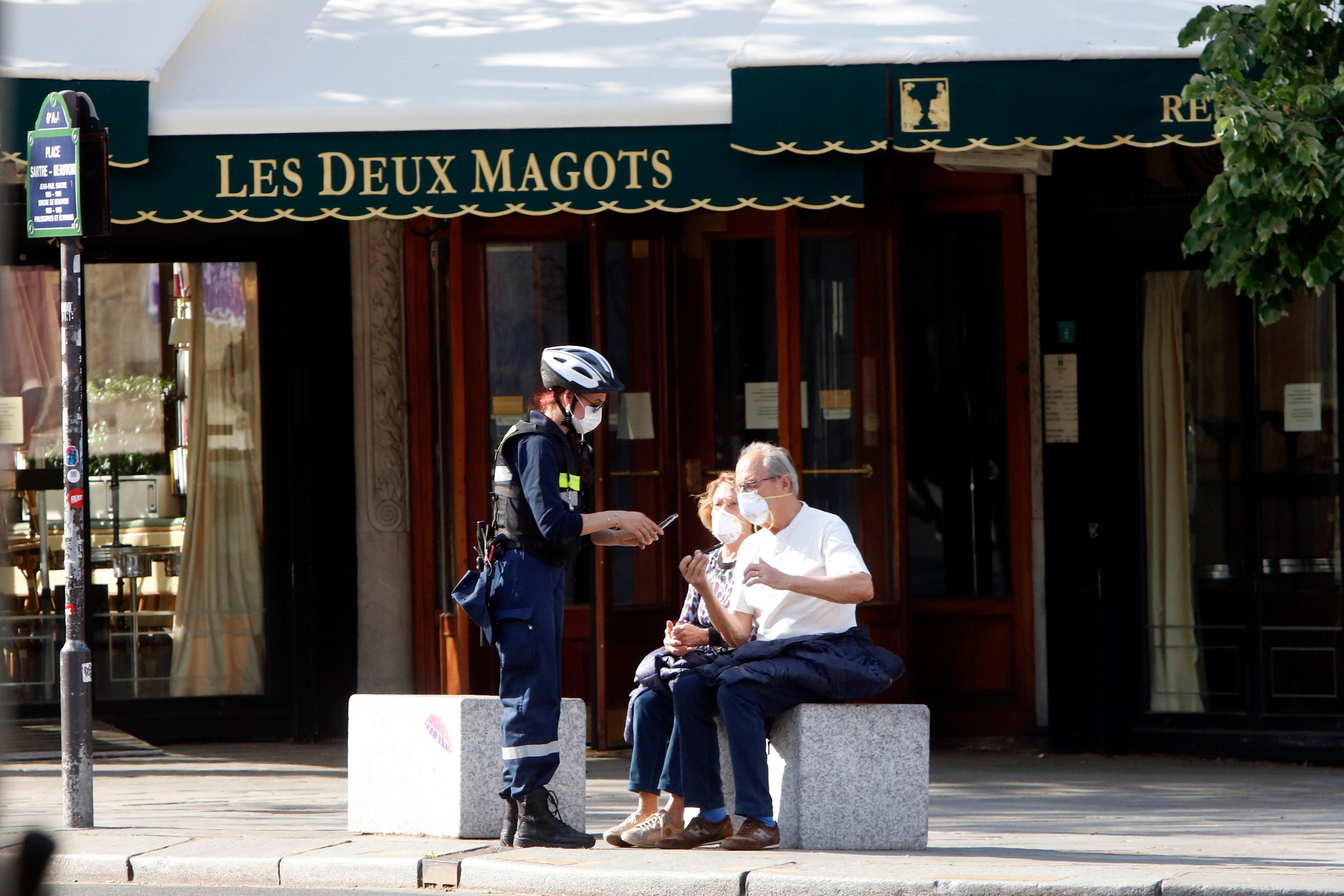 Photo of a masked officer talking to two seated people outside Les Deux Magots cafe in Paris.