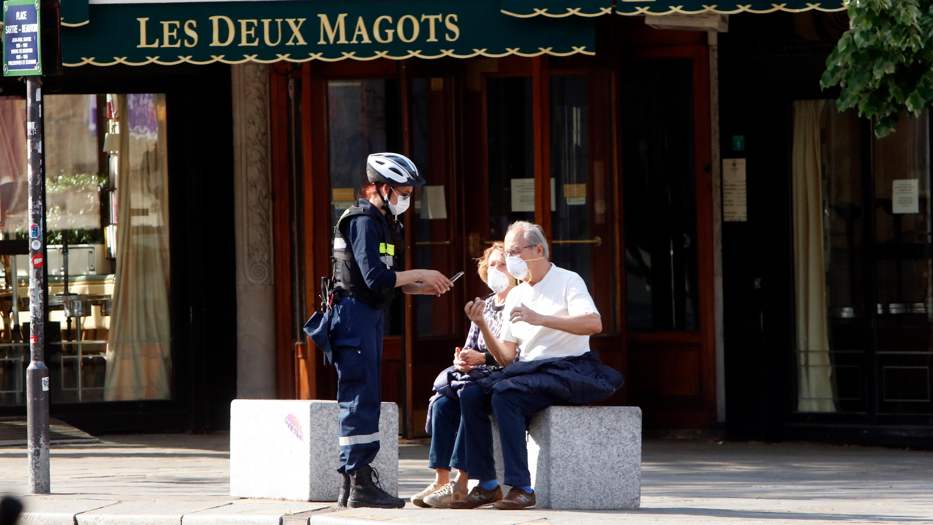 Photo of a masked officer talking to two seated people outside Les Deux Magots cafe in Paris.
