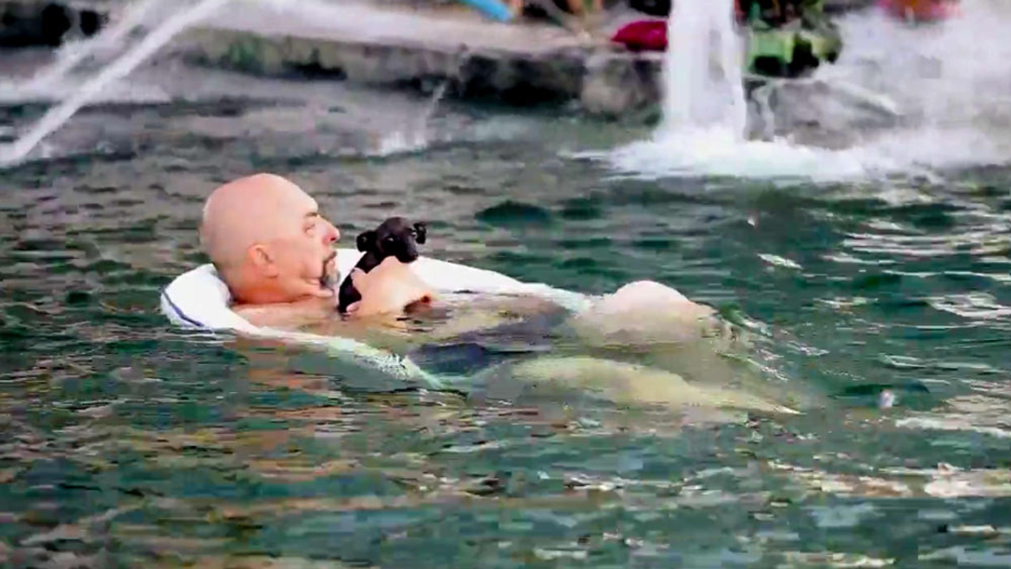 A bald man floating on his back in a pool while holding a small black puppy. Water fountains are in the background.