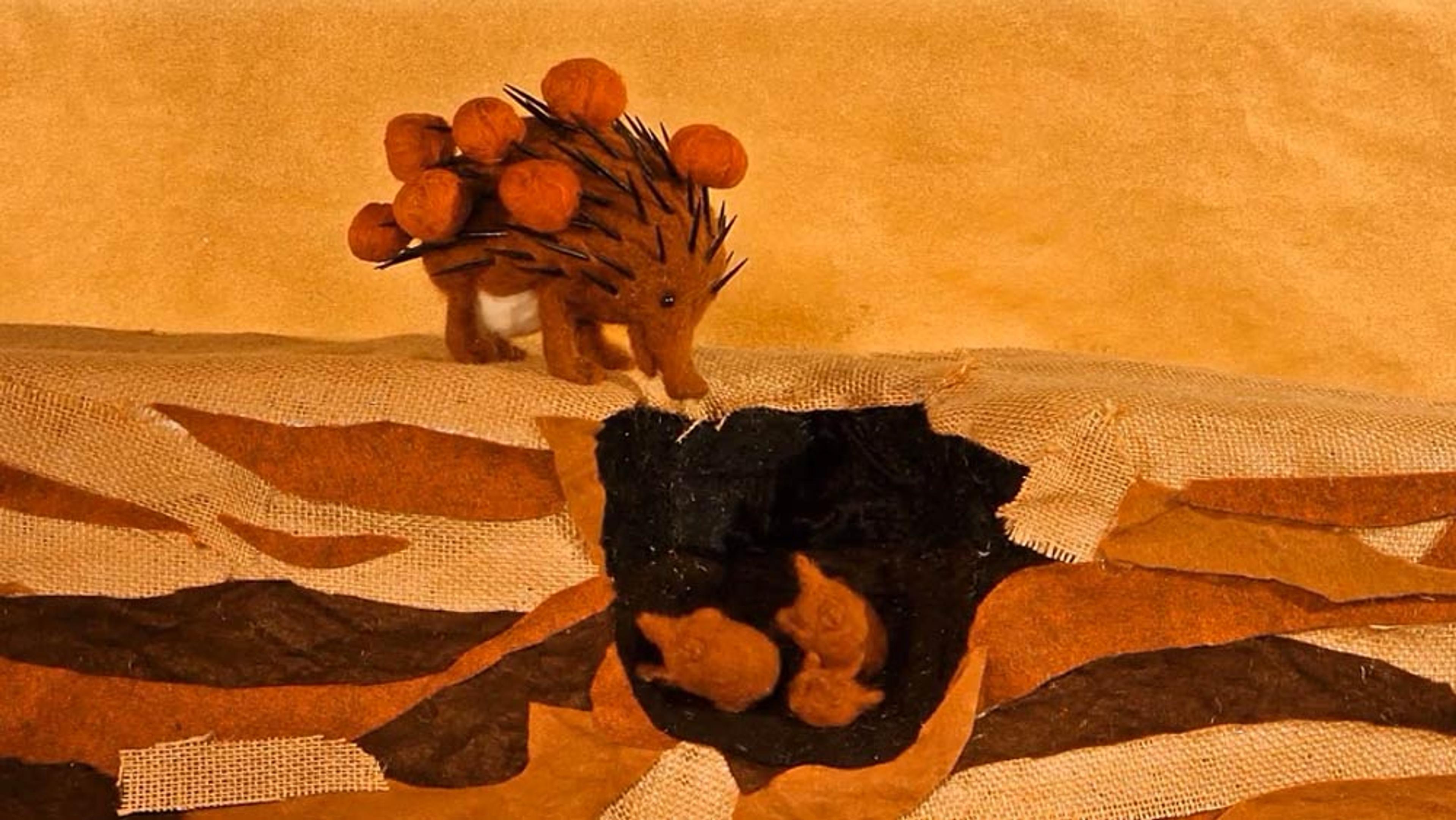 A felt art scene with a hedgehog carrying mushrooms above a burrow with two small animals inside. Background in earthy tones.