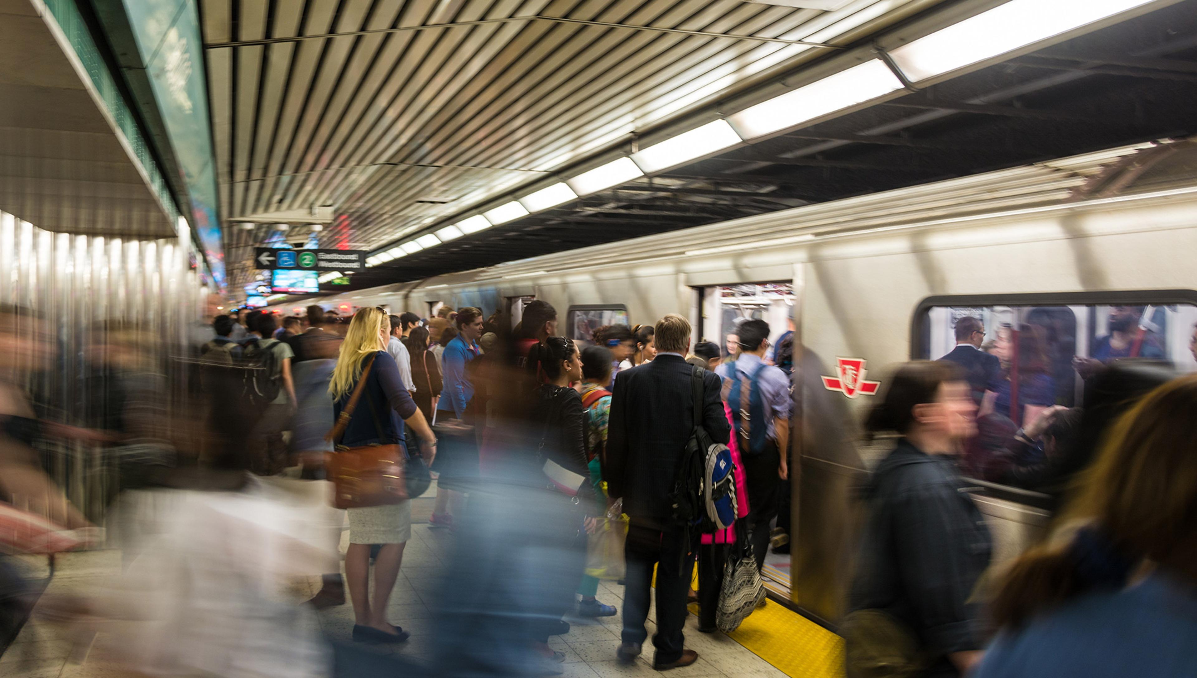 A train arrives at a busy subway station. Many different people on the platform wait to get on the train. Some are blurred as they are caught by the camera mid-motion.