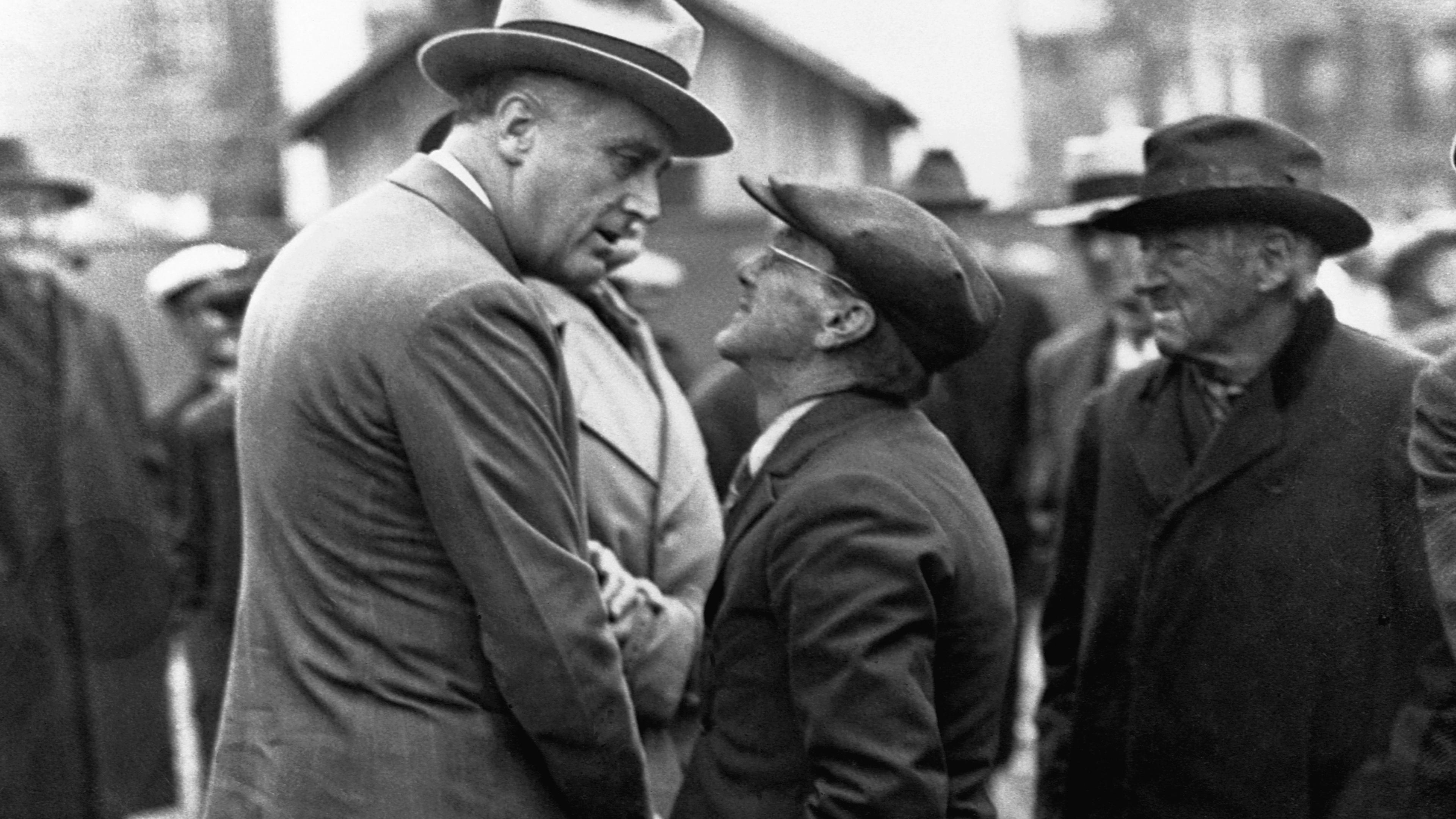 Black-and-white photo of two men in hats conversing on a street, one very tall, the other short, with onlookers nearby.