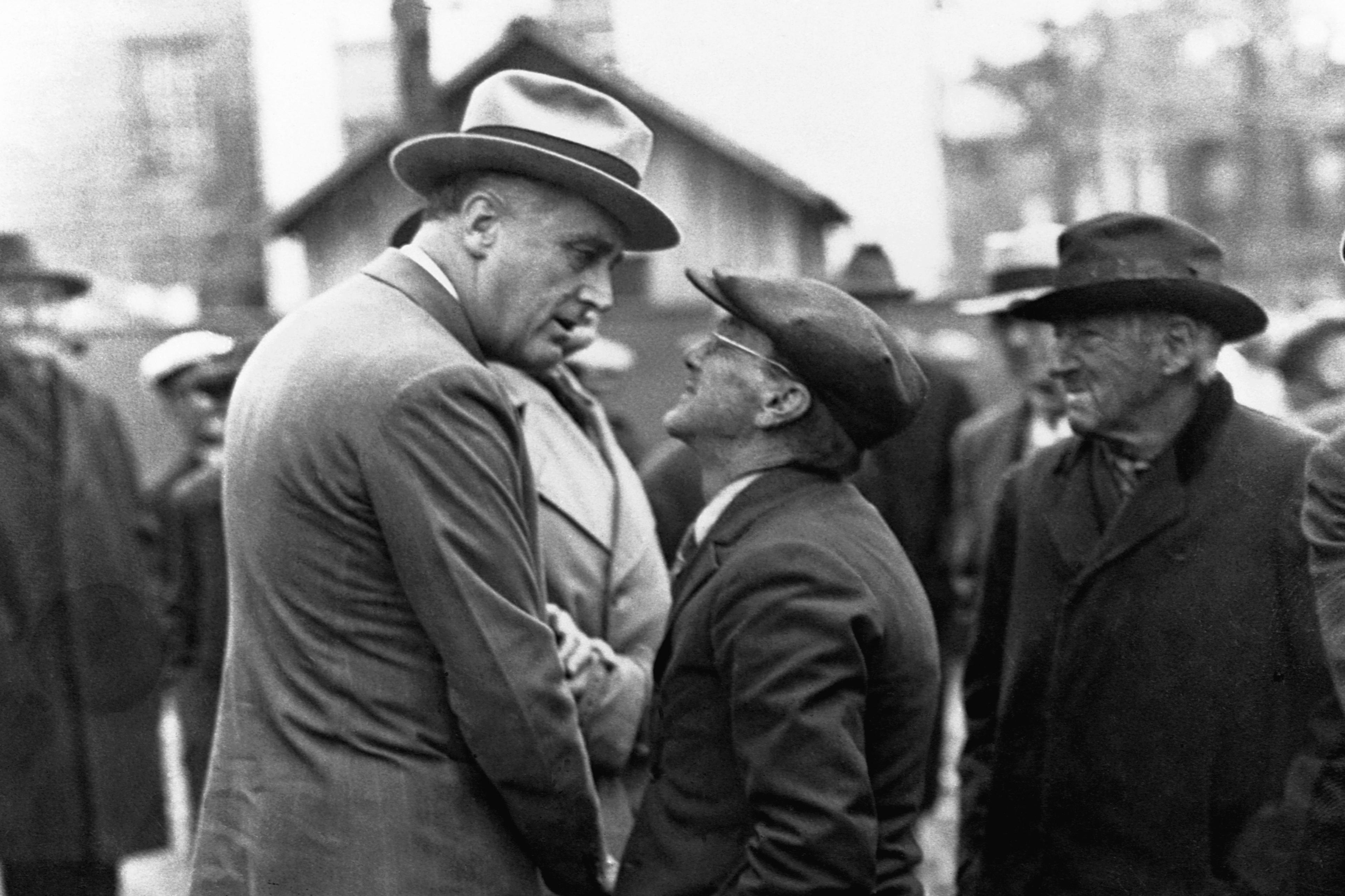 Black-and-white photo of two men in hats conversing on a street, one very tall, the other short, with onlookers nearby.