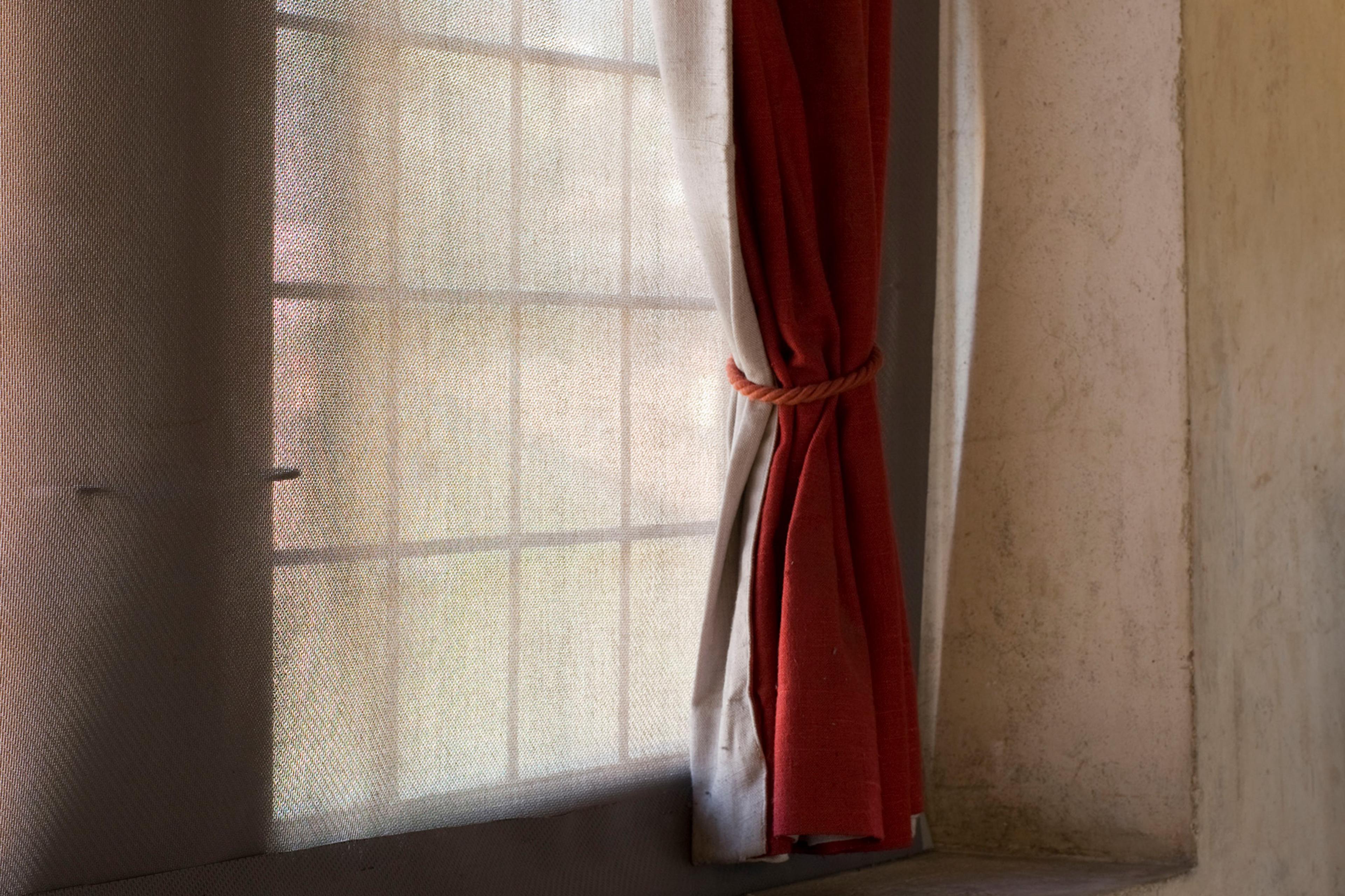 A window with light streaming through sheer curtains, partially covered by a red curtain tied back with a rope tie.