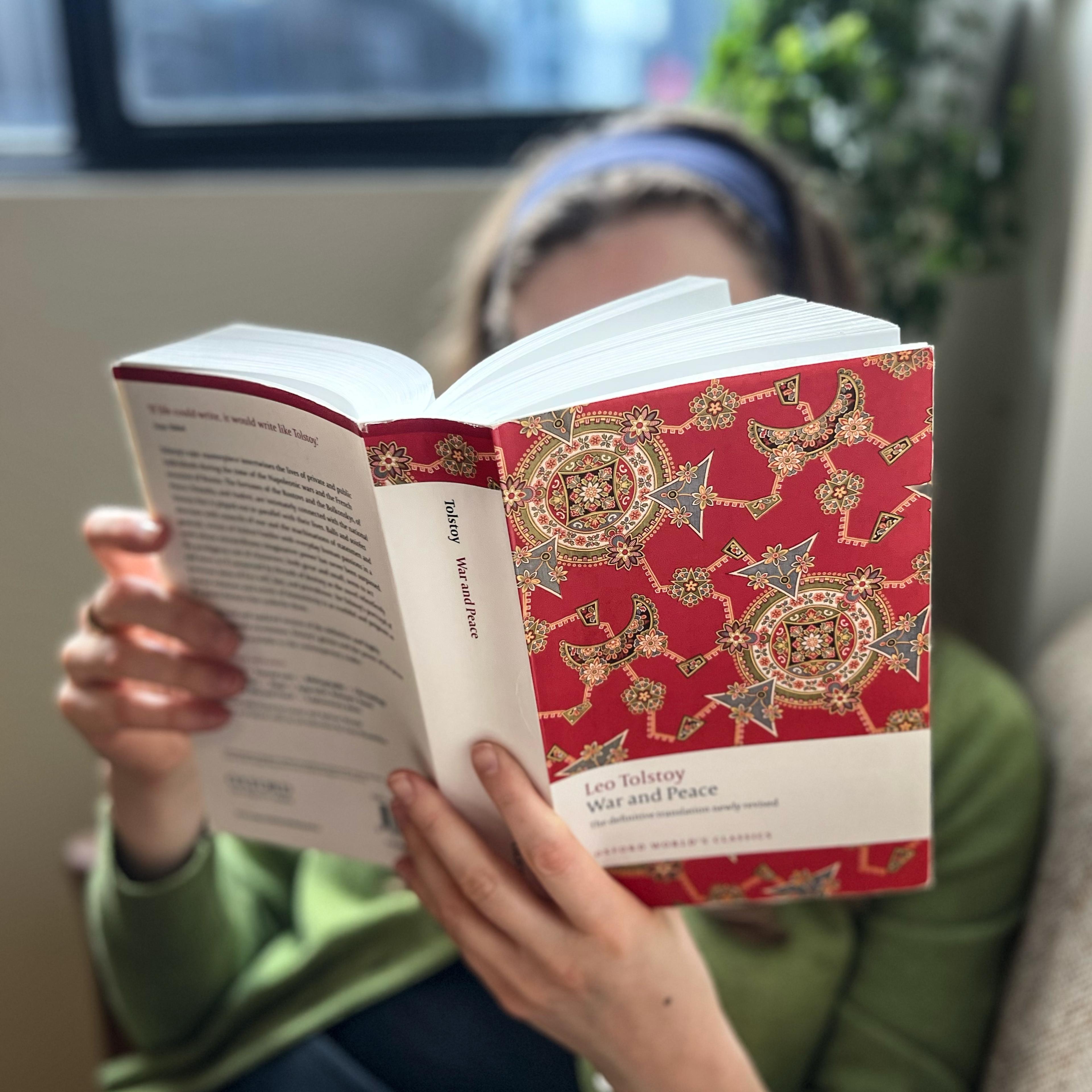 창문 가까이에 앉아 붉은 장식 표지에 톨스토이의 '전쟁과 평화'를 읽고 있는 사람의 사진입니다.<BR>