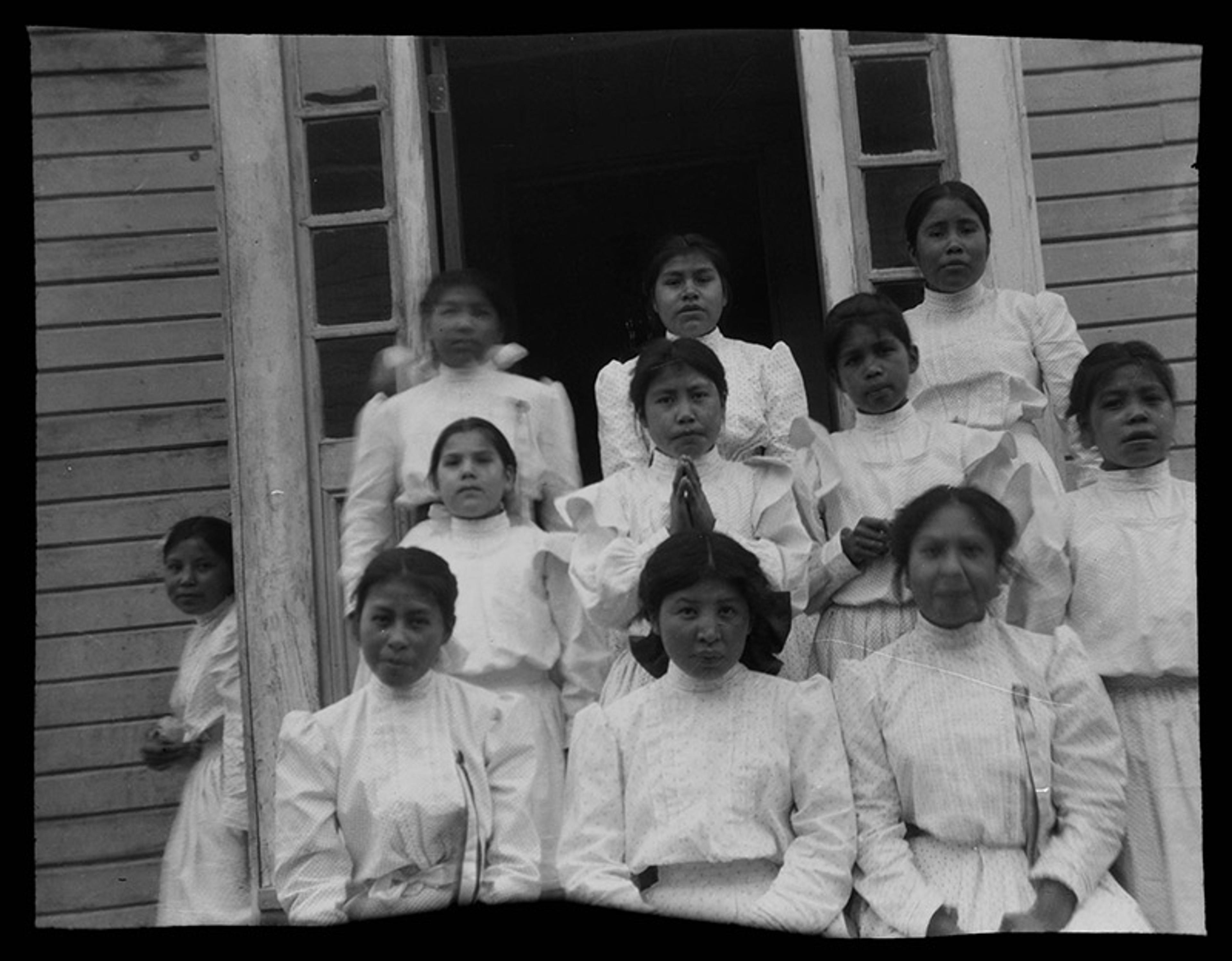 Black and white photo of a group of young girls in white dresses standing on steps in front of a building, likely a school or institution.