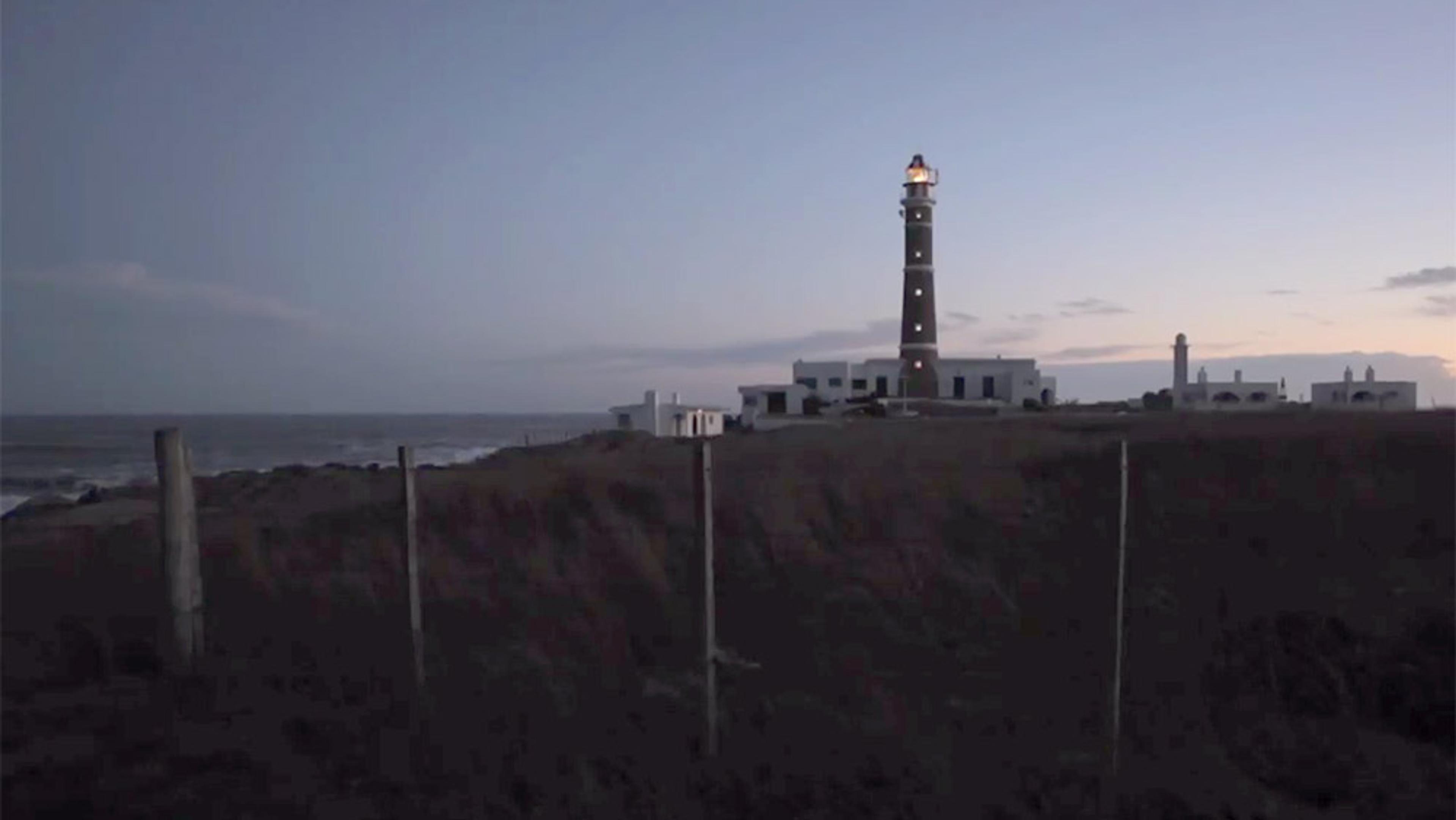 A lit lighthouse at dusk next to white buildings on a coastline with a fence in the foreground and the sea on the left.
