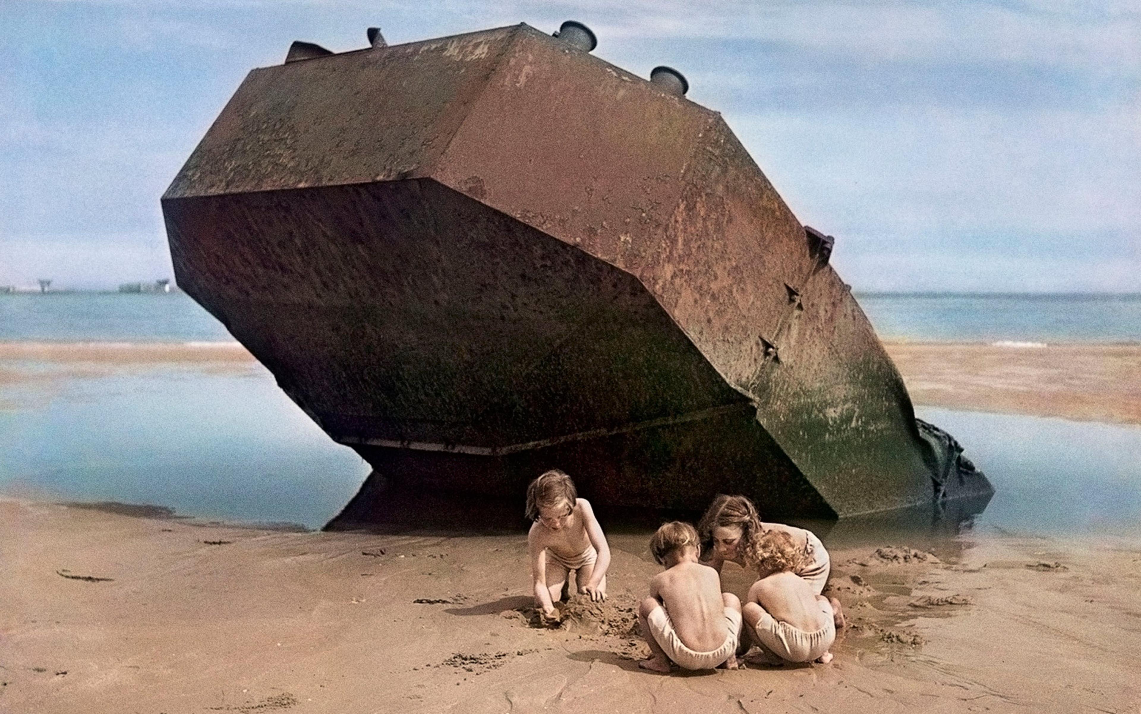 Four small children playing in the shadow of a large rusted military boat beached in the sand with the sea in the background.
