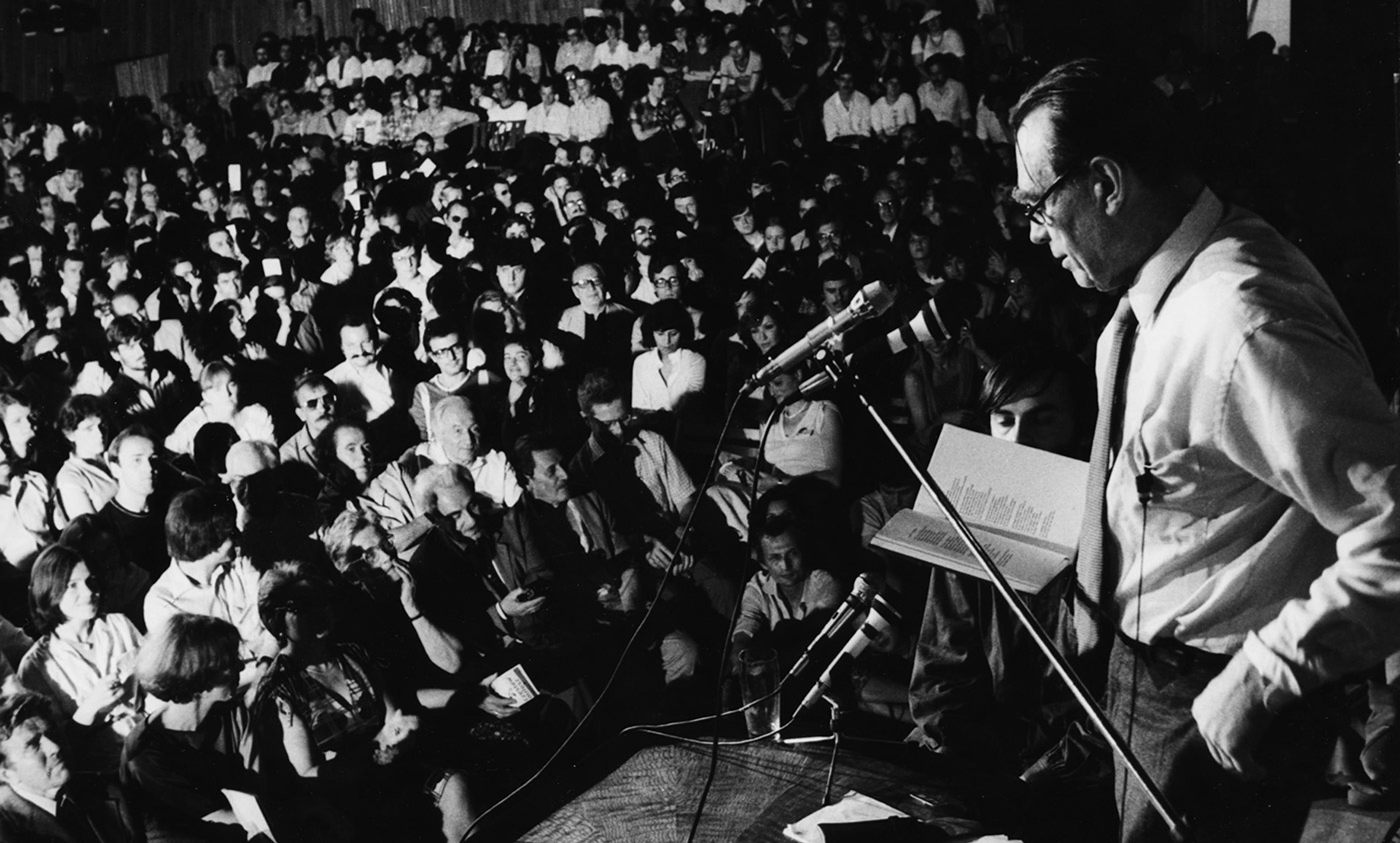 Black and white photo of a man speaking into a microphone in front of a large audience sitting in close rows attentively listening.