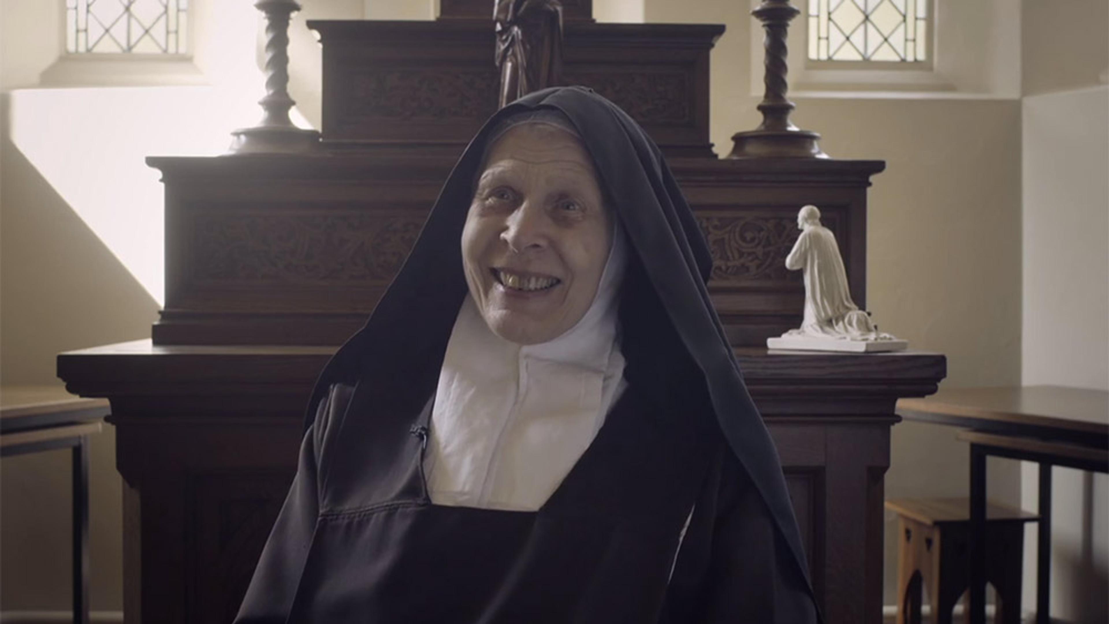 A smiling nun in a black habit and white wimple sitting in a church, with wooden furniture and a statue in the background.