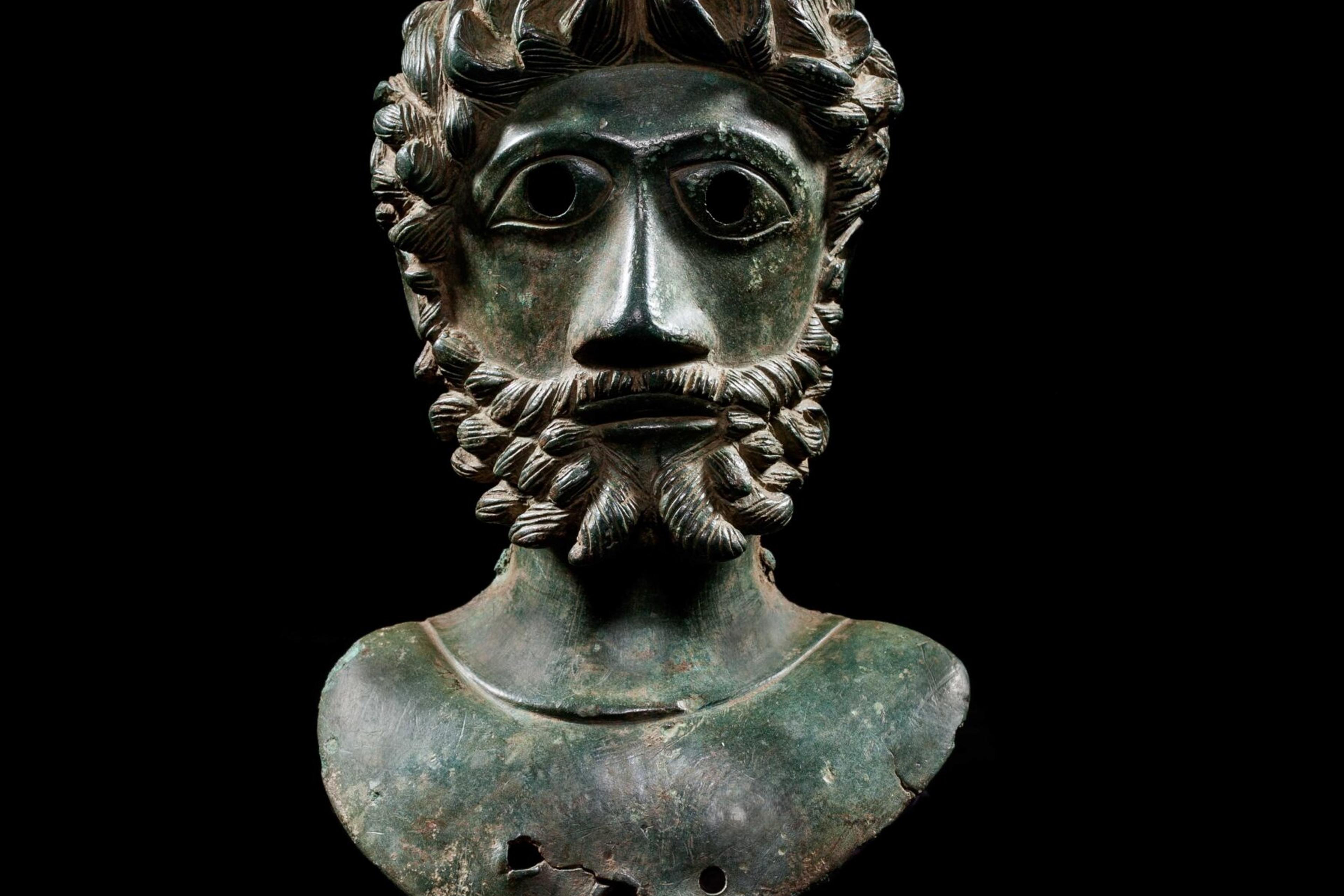 Bronze bust of a bearded man with curled hair against a black background, ancient artefact.