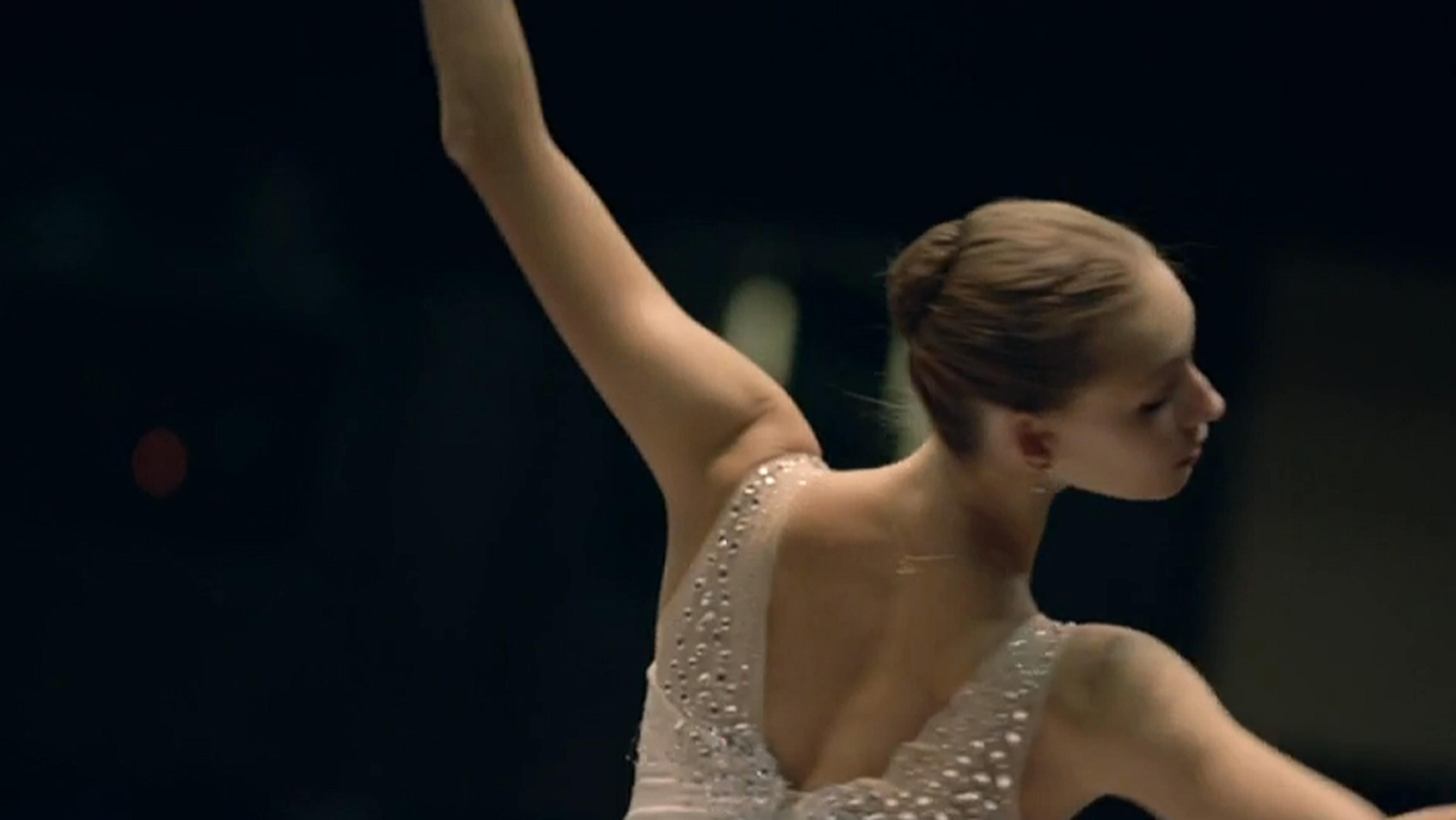 A ballet dancer in a white costume with beads, arm raised, hair in a bun, captured from the back on a dark background.