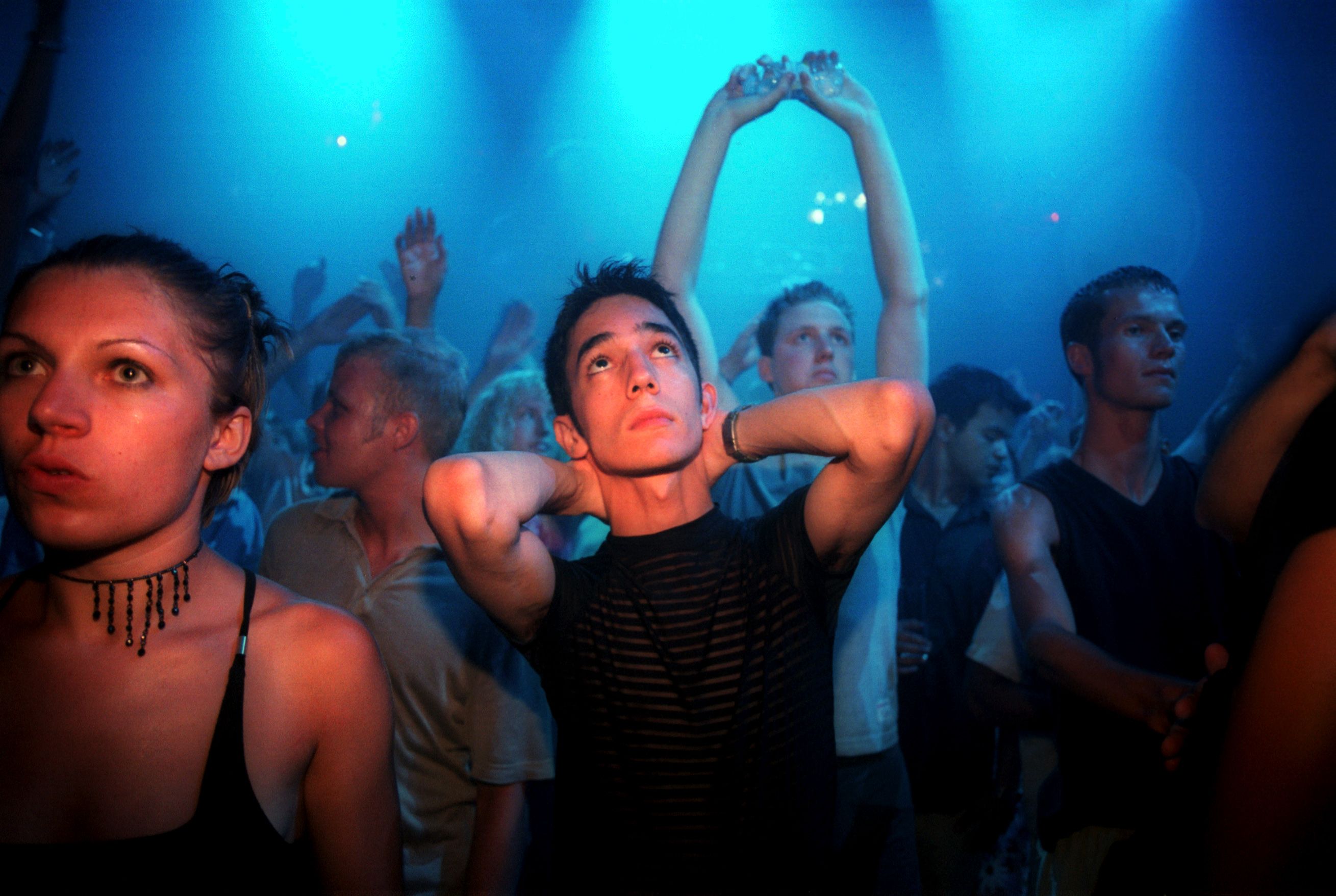 A crowded dance club with blue lighting people dancing some looking upwards hands raised in the air expressions vary.