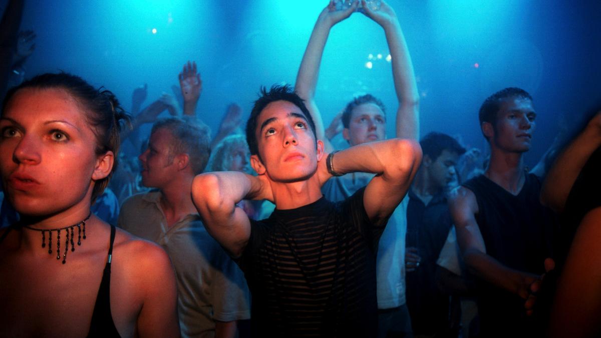 A crowded dance club with blue lighting people dancing some looking upwards hands raised in the air expressions vary.