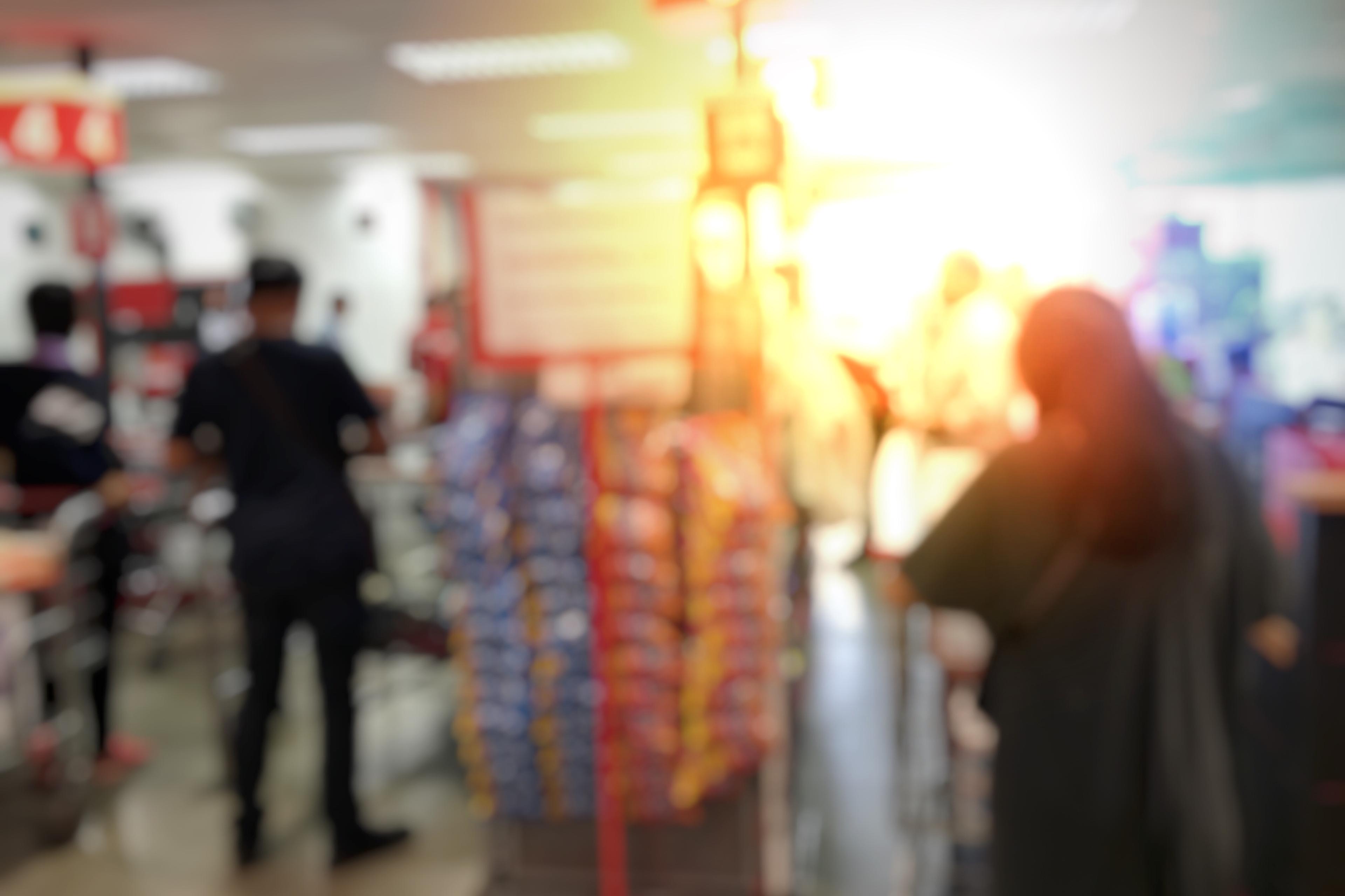 Blurry photo of a supermarket interior with people shopping, bright lighting and shelves filled with products.