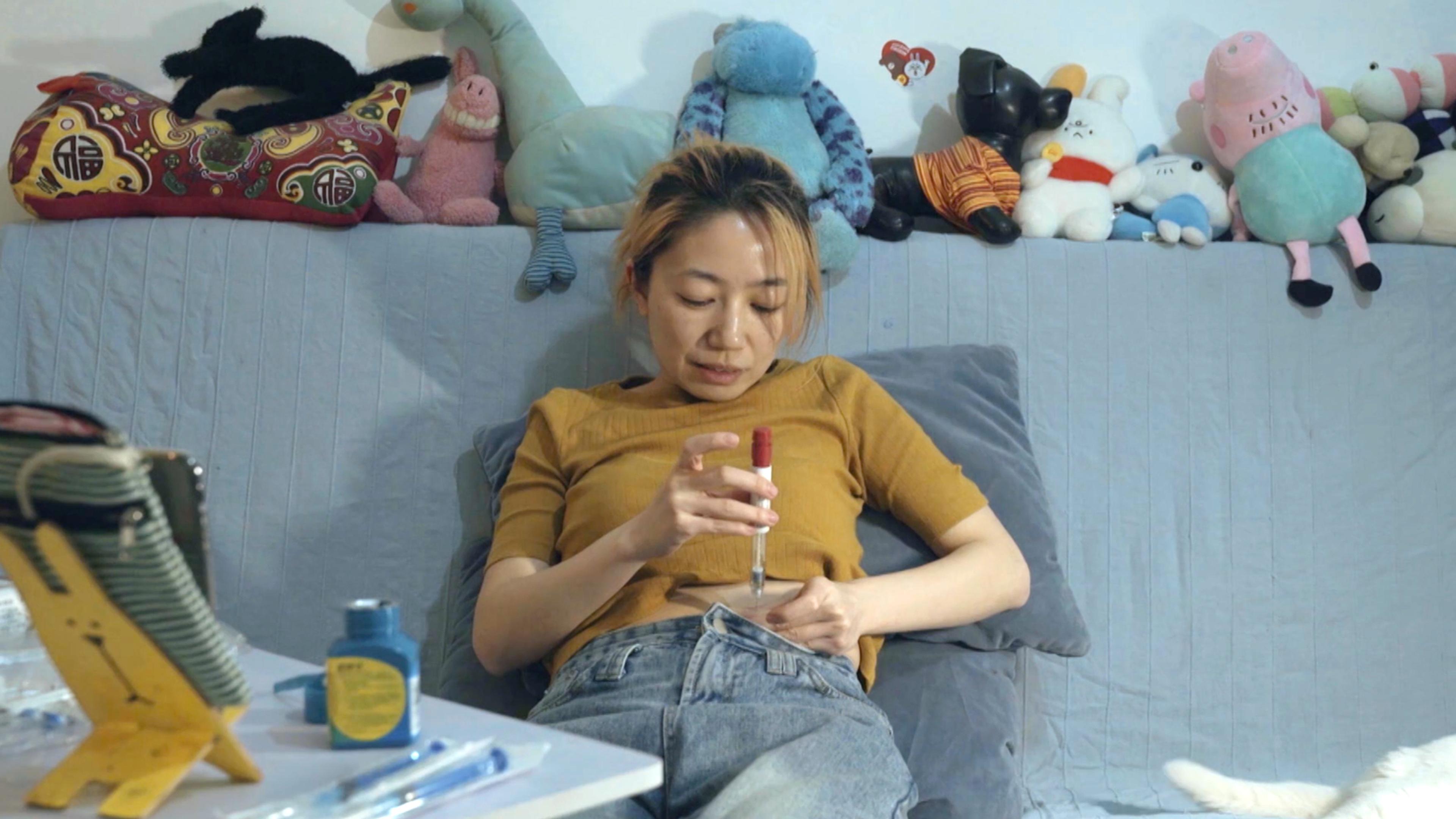 A woman on a sofa injecting her abdomen surrounded by stuffed toys on a shelf.