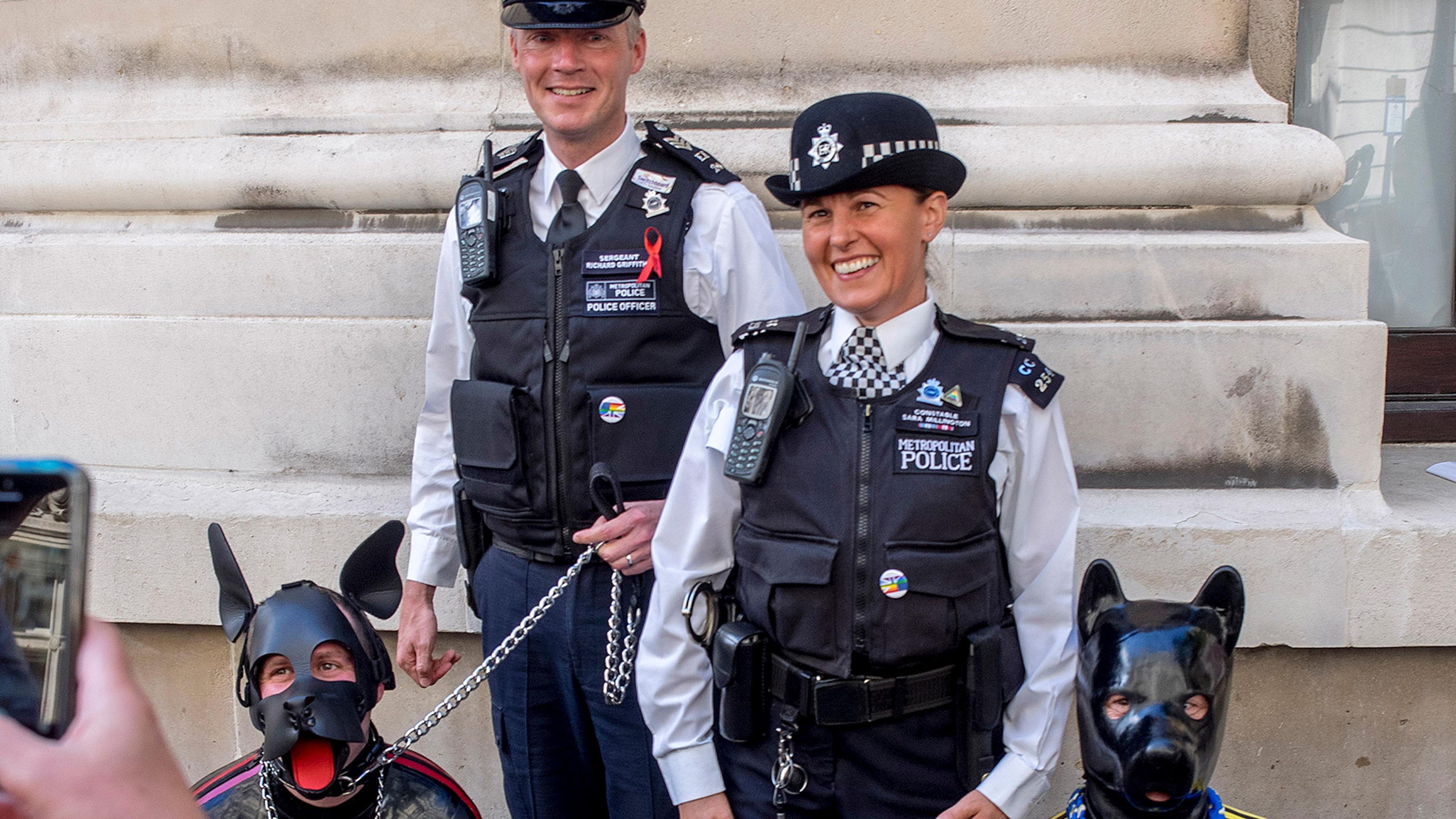 Photo of two police officers smiling next to two people in dog costumes, with one person capturing the moment on a phone.