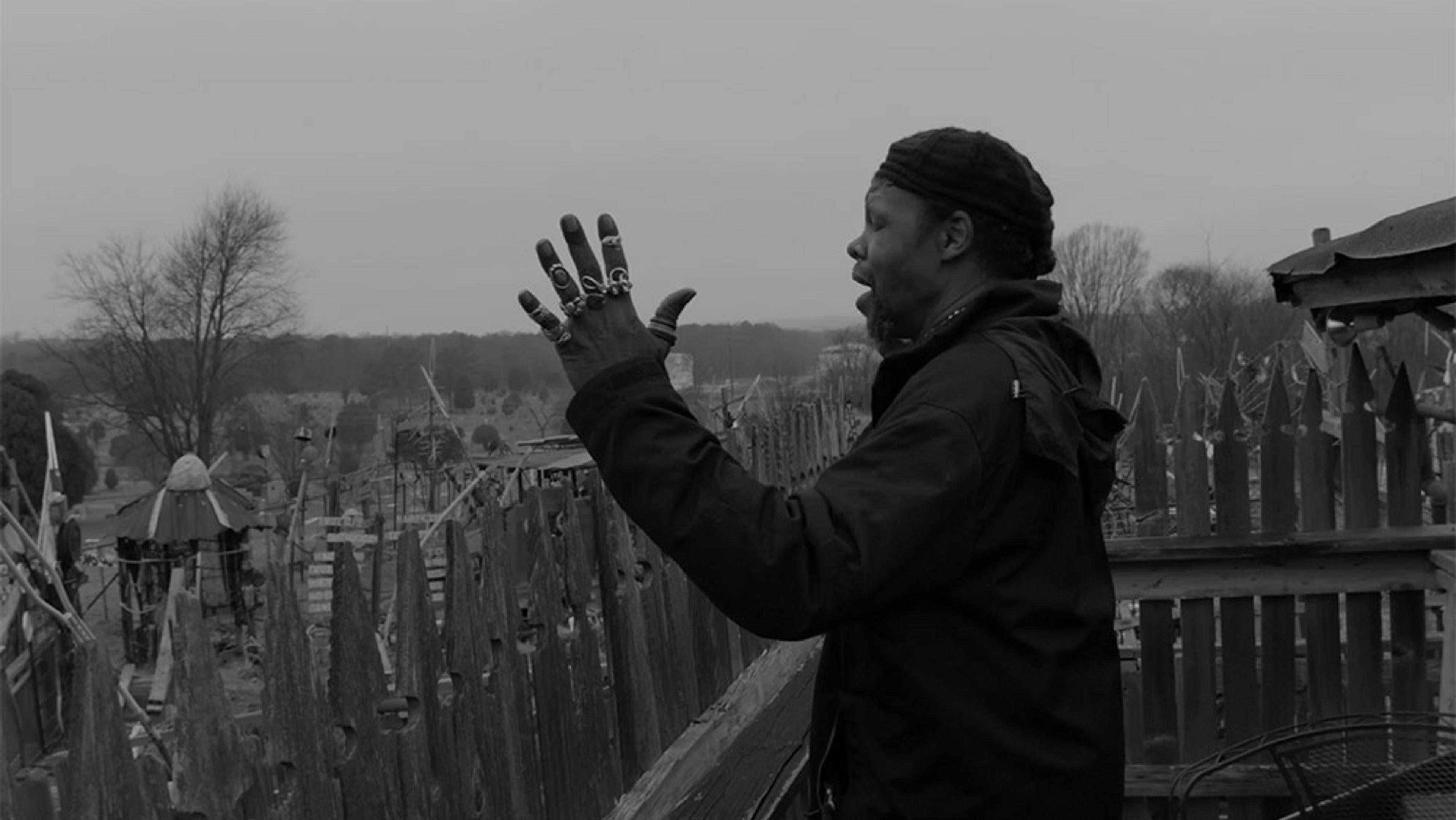 Black and white photo of a Black man with rings on his hands talking with eyes closed beside a wooden fence with trees and a cloudy sky in the background.