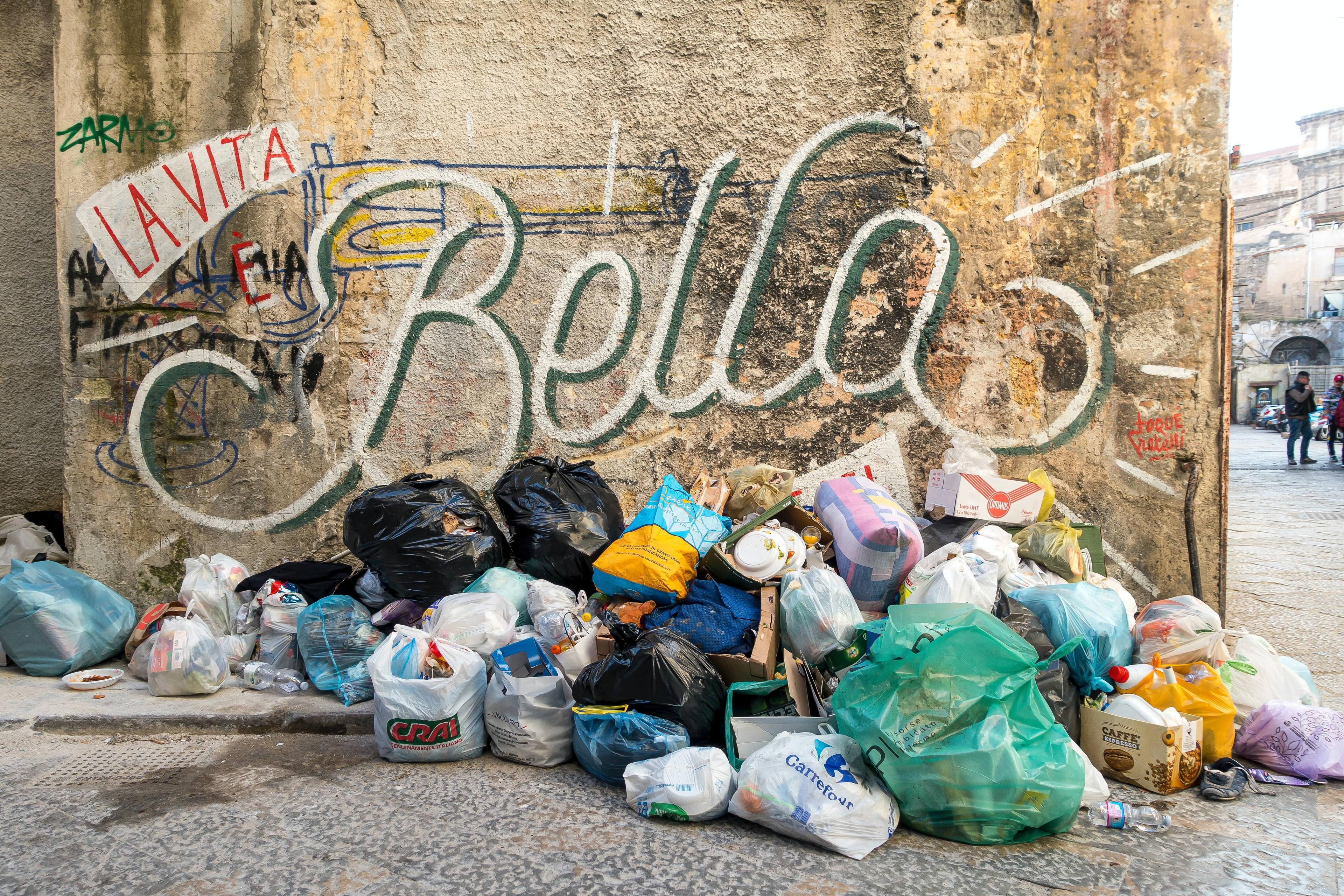 Photo of rubbish piled against a wall with graffiti reading “La Vita è Bella” on a city street.