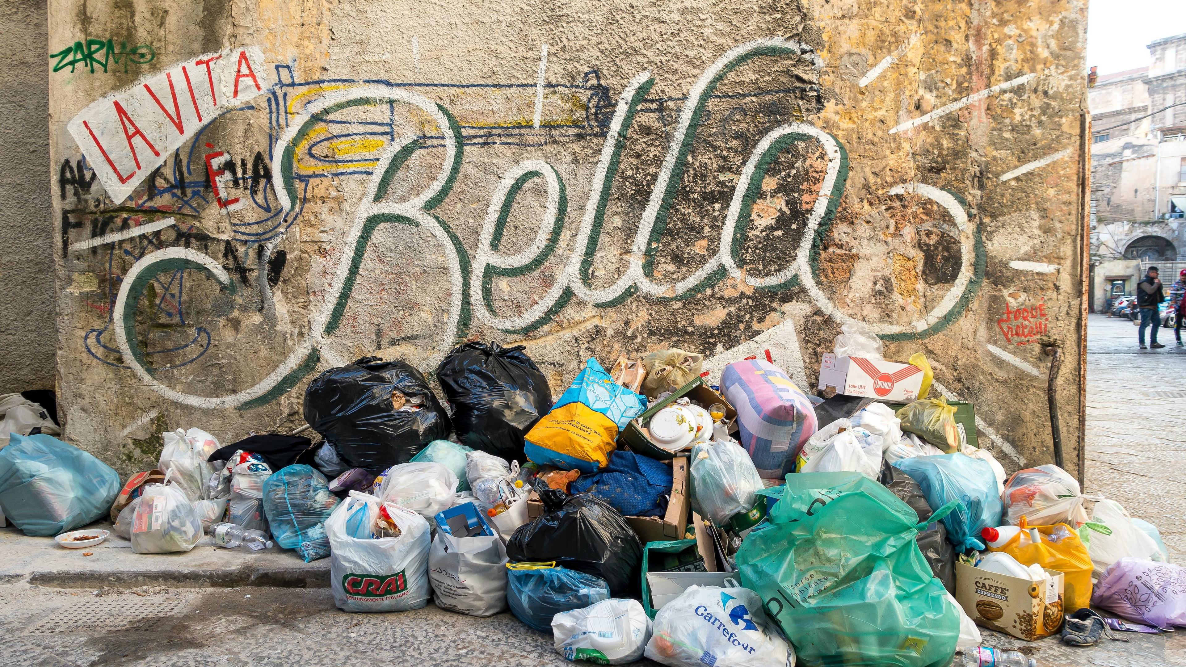 Photo of rubbish piled against a wall with graffiti reading “La Vita è Bella” on a city street.