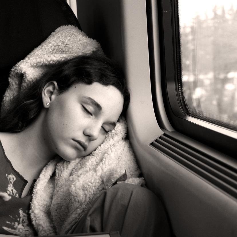 Black-and-white photo of a person sleeping against a window on a train, head resting on a pillow, outside view blurred.