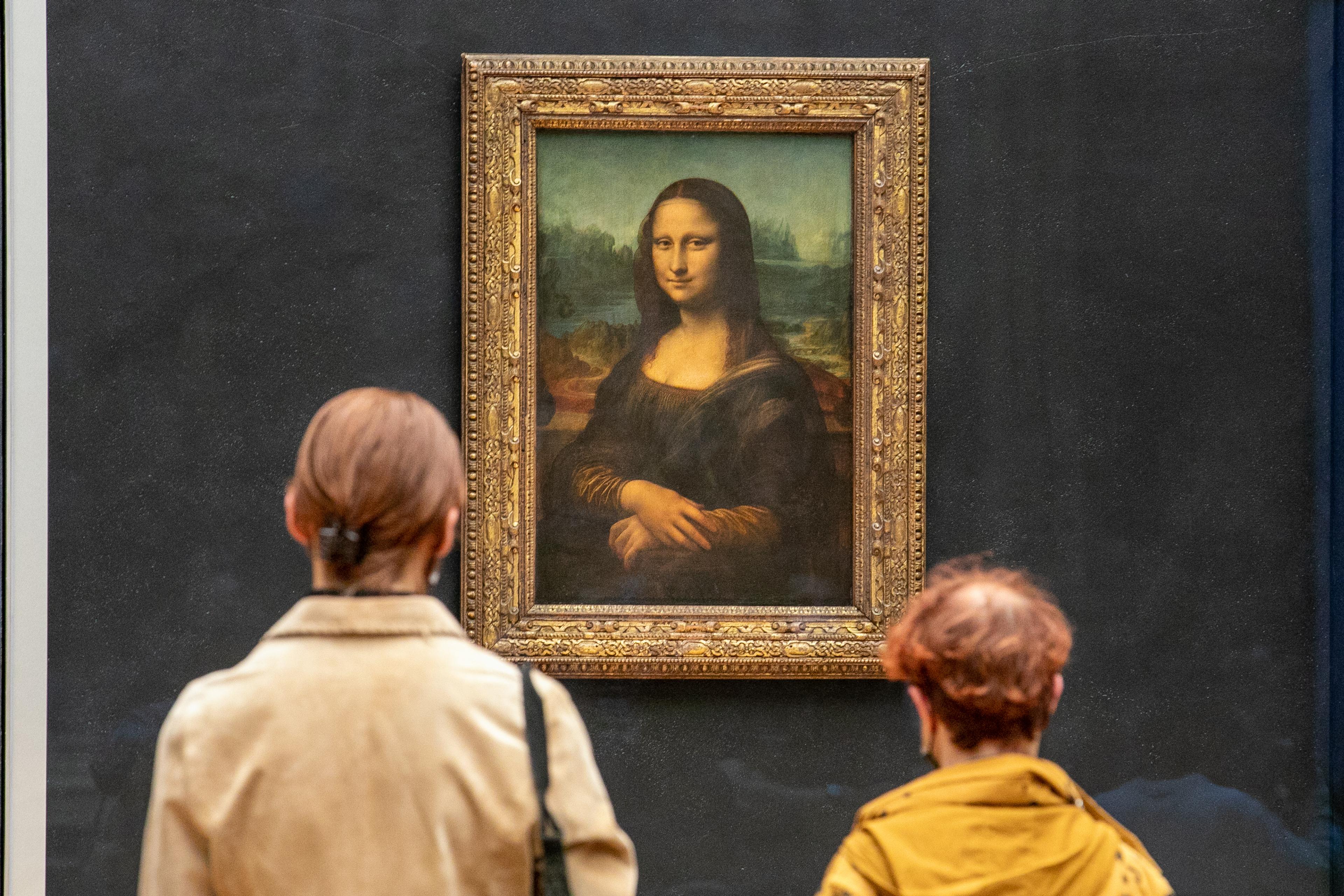 Two people view a famous painting in a gallery; it’s the Mona Lisa in an ornate frame.