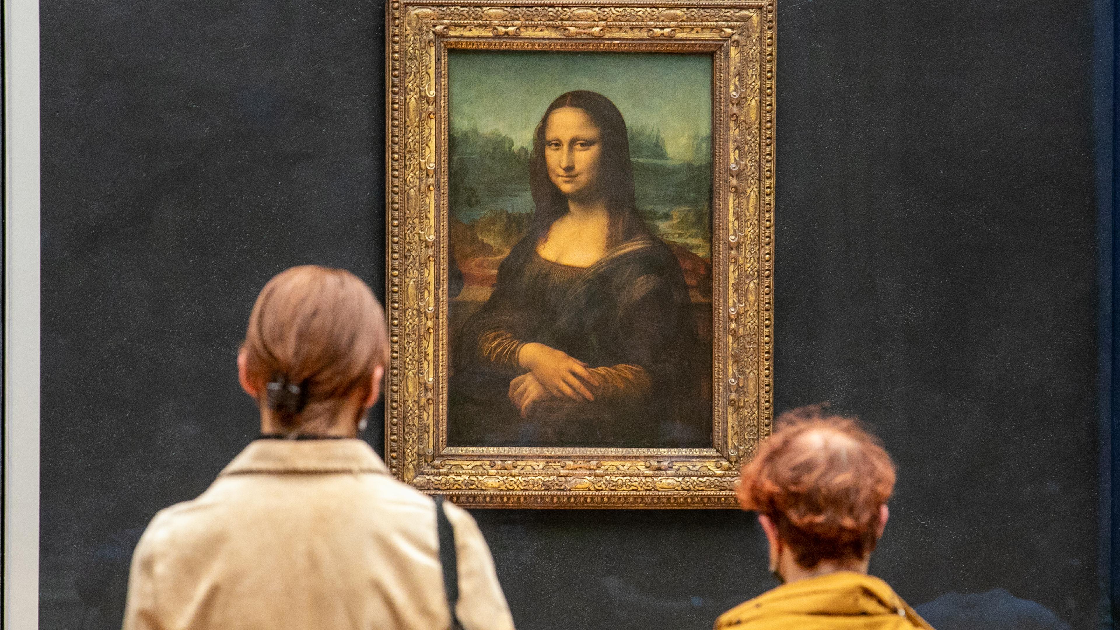 Two people view a famous painting in a gallery; it’s the Mona Lisa in an ornate frame.