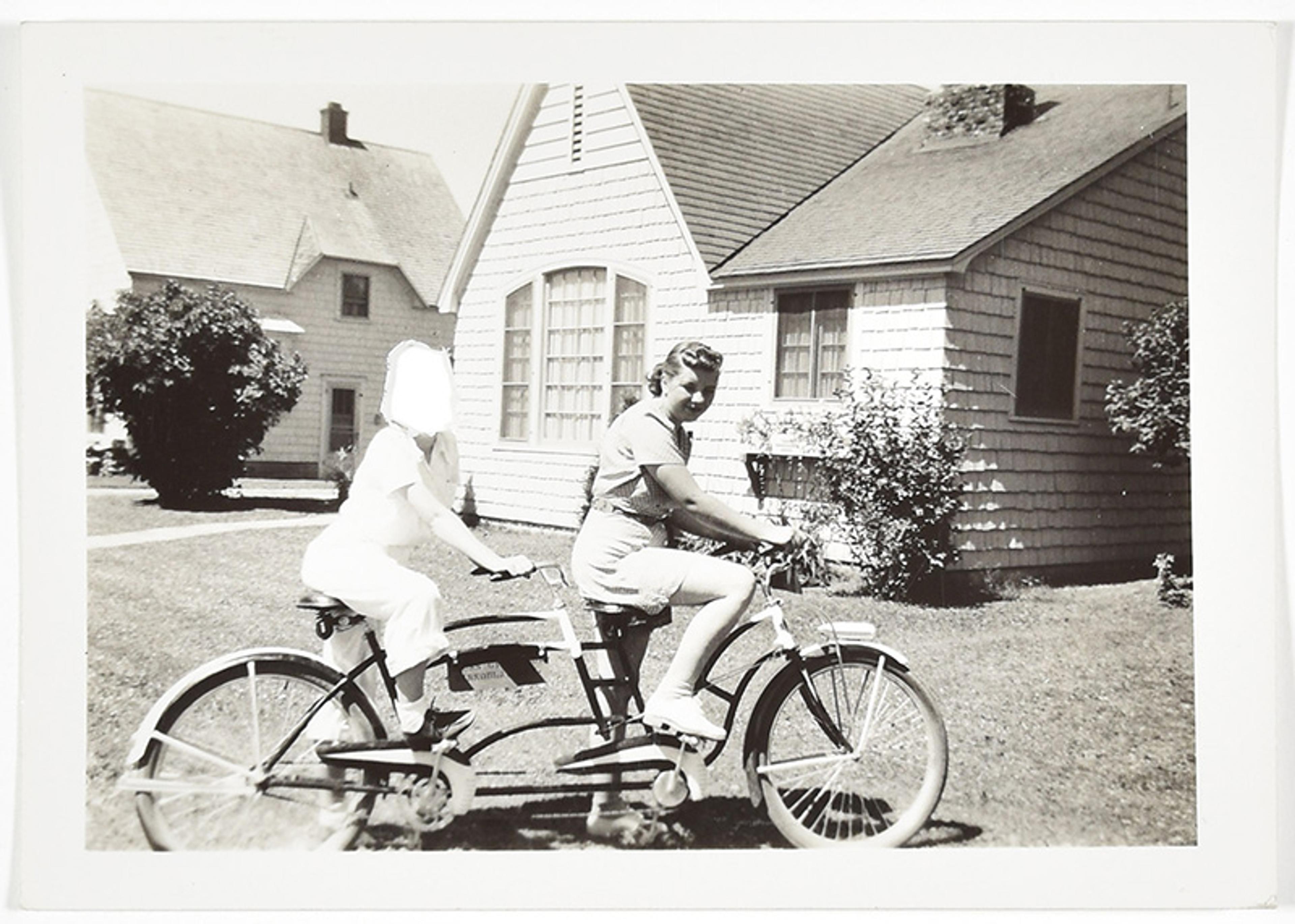 Vintage photo of two women, one with her face obscured, on a tandem bicycle in front of a shingled house with a garden on a sunny day.