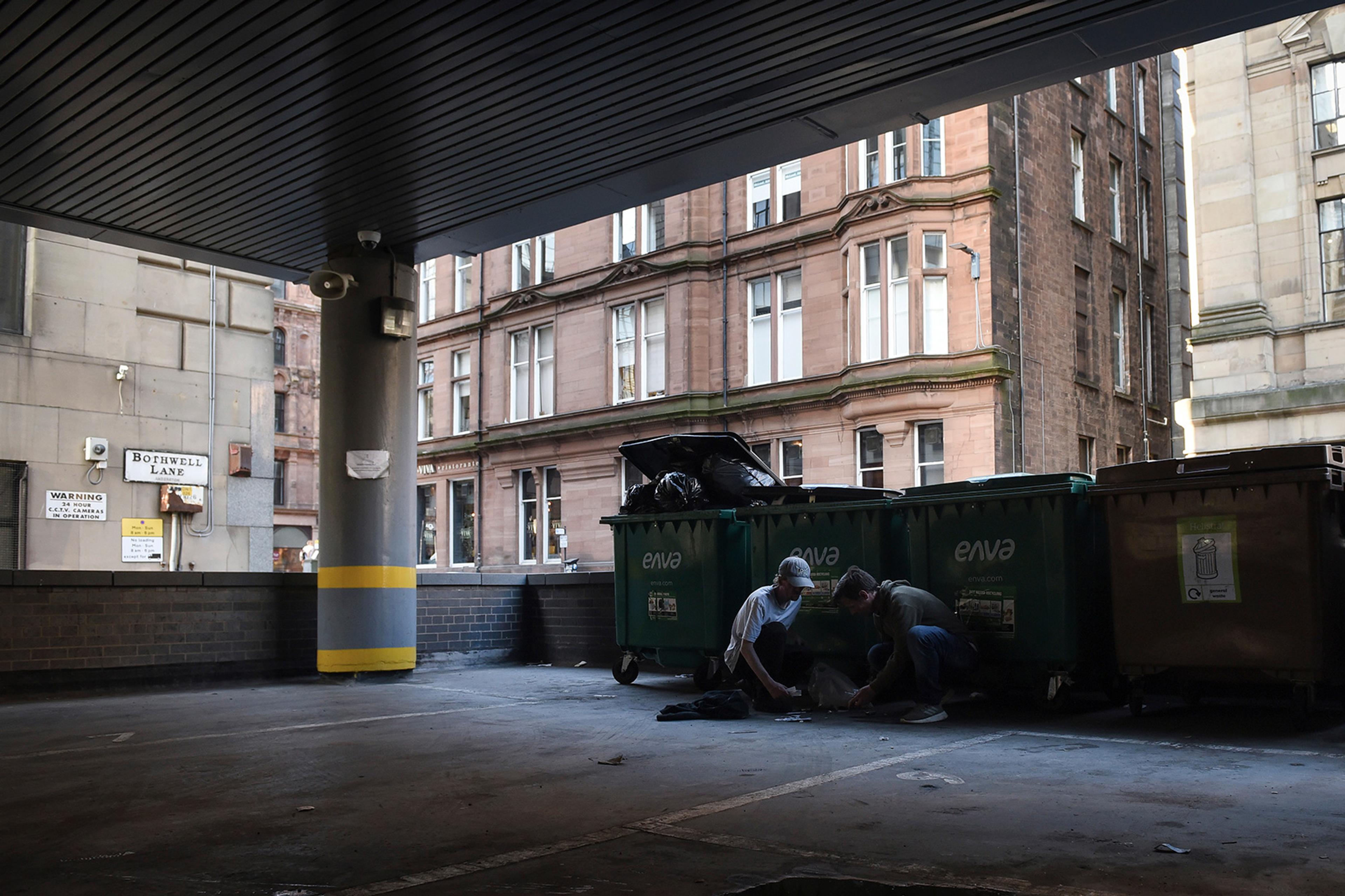 Two people squatting next to large bins in an alley named Bothwell Lane, with old brick buildings in the background.