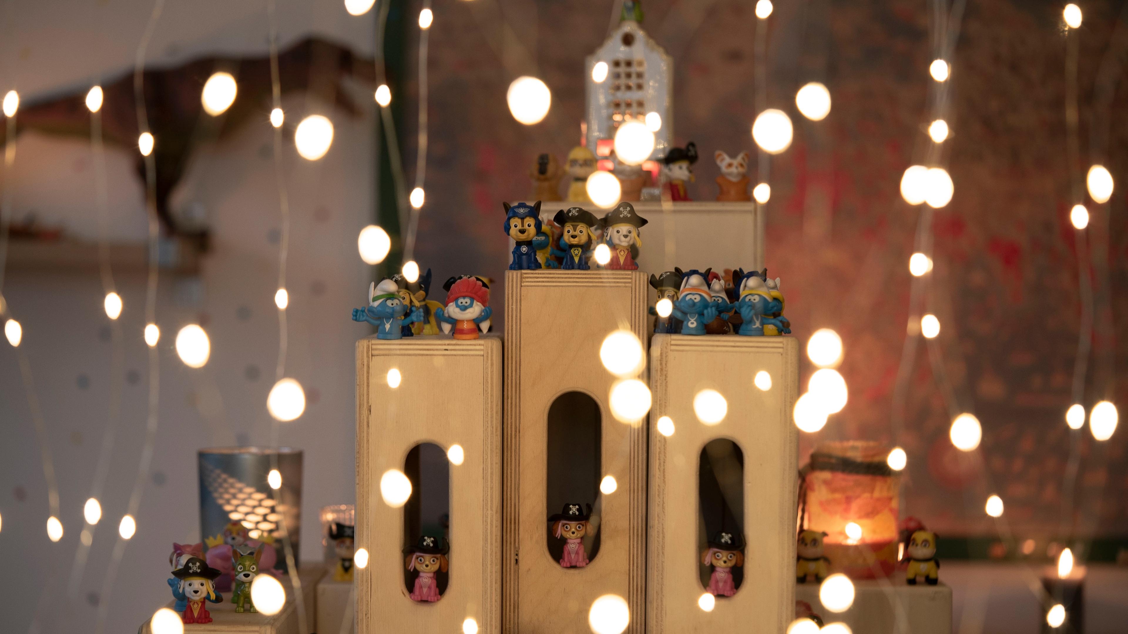 Photo of miniature figurines on wooden blocks with strings of glowing lights in the foreground creating a festive atmosphere.