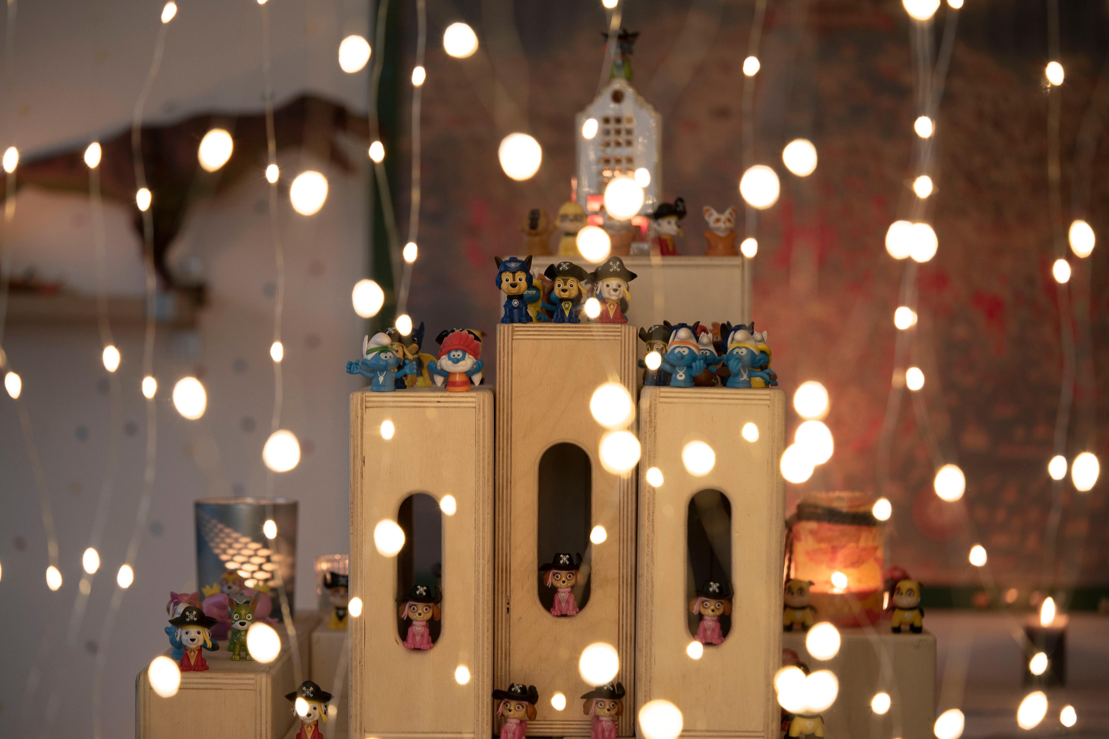 Photo of miniature figurines on wooden blocks with strings of glowing lights in the foreground creating a festive atmosphere.