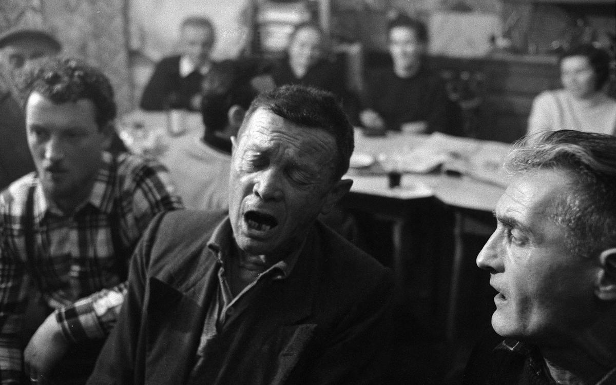 A black-and-white photo of a group of men sitting close together, some with mouths open as if singing or speaking. In the blurry background, additional people are seated around a table. The scene appears to be inside a room with a warm, communal atmosphere.