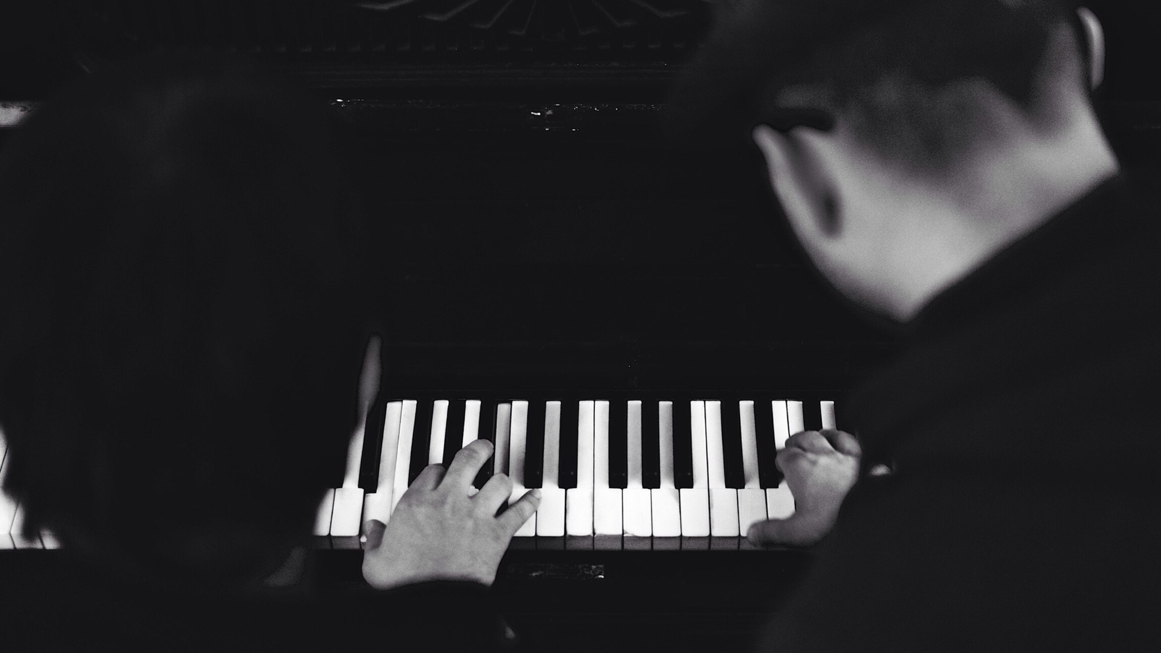 Black and white photo of two people playing a piano, focused on their hands and the keyboard from above.