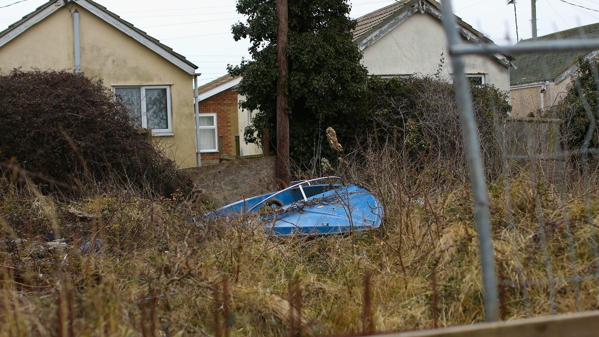An overgrown garden with a blue boat surrounded by tall grass, houses in the background and a wooden fence in the foreground.