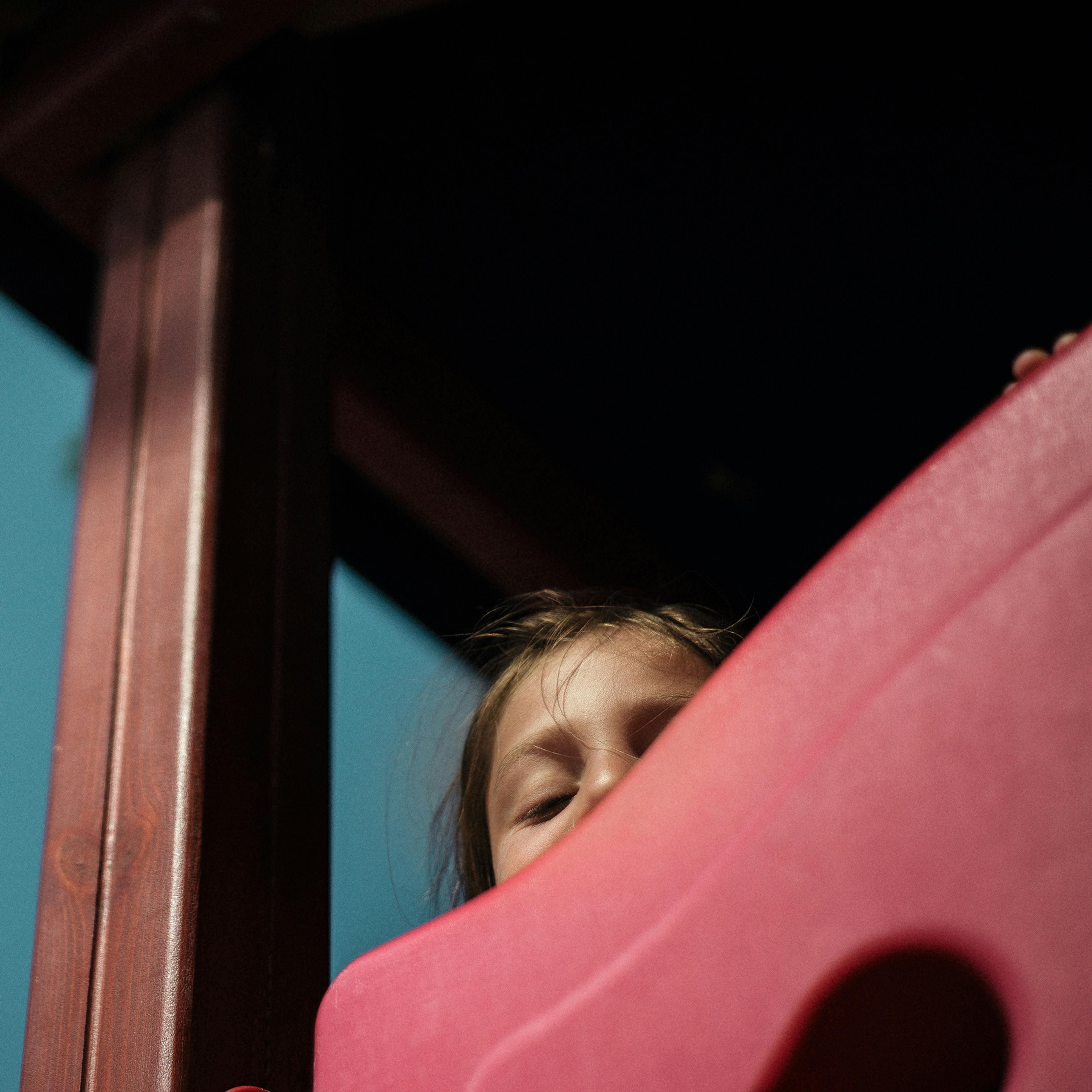 a frightened child whose face is partially obscured peers down from by a play area structure
