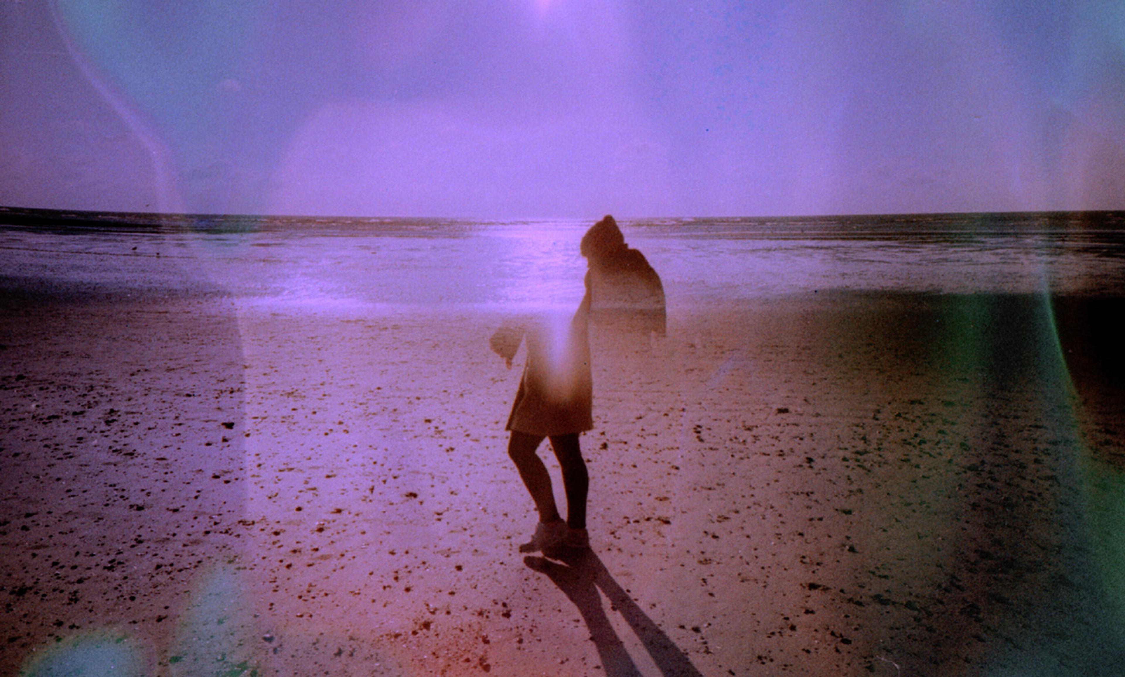 A person on a beach at sunset, with light leaks and a purple hue casting a long shadow on the sand.