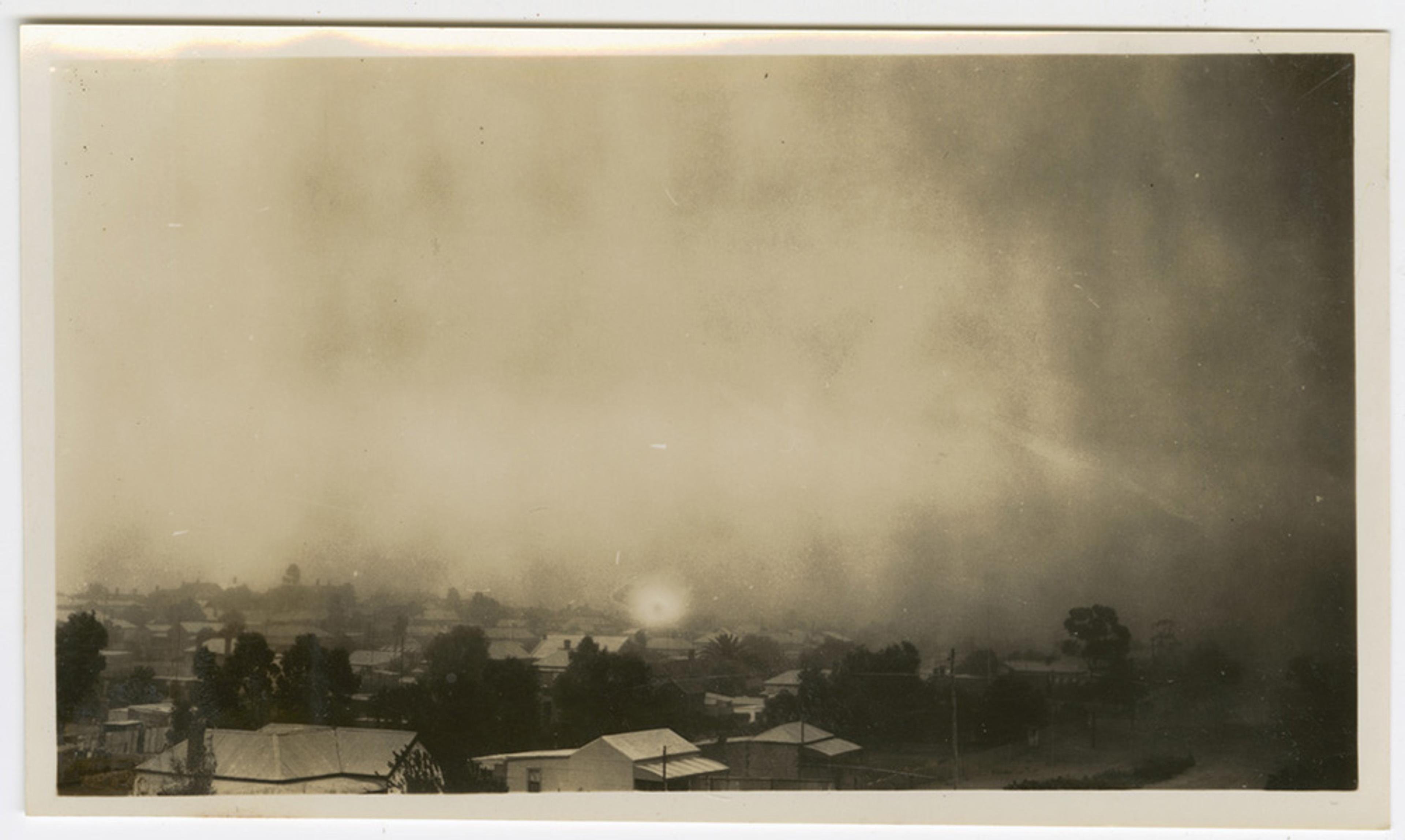 Black and white photo of a town shrouded in thick fog or dust, buildings and trees barely visible through the haze.