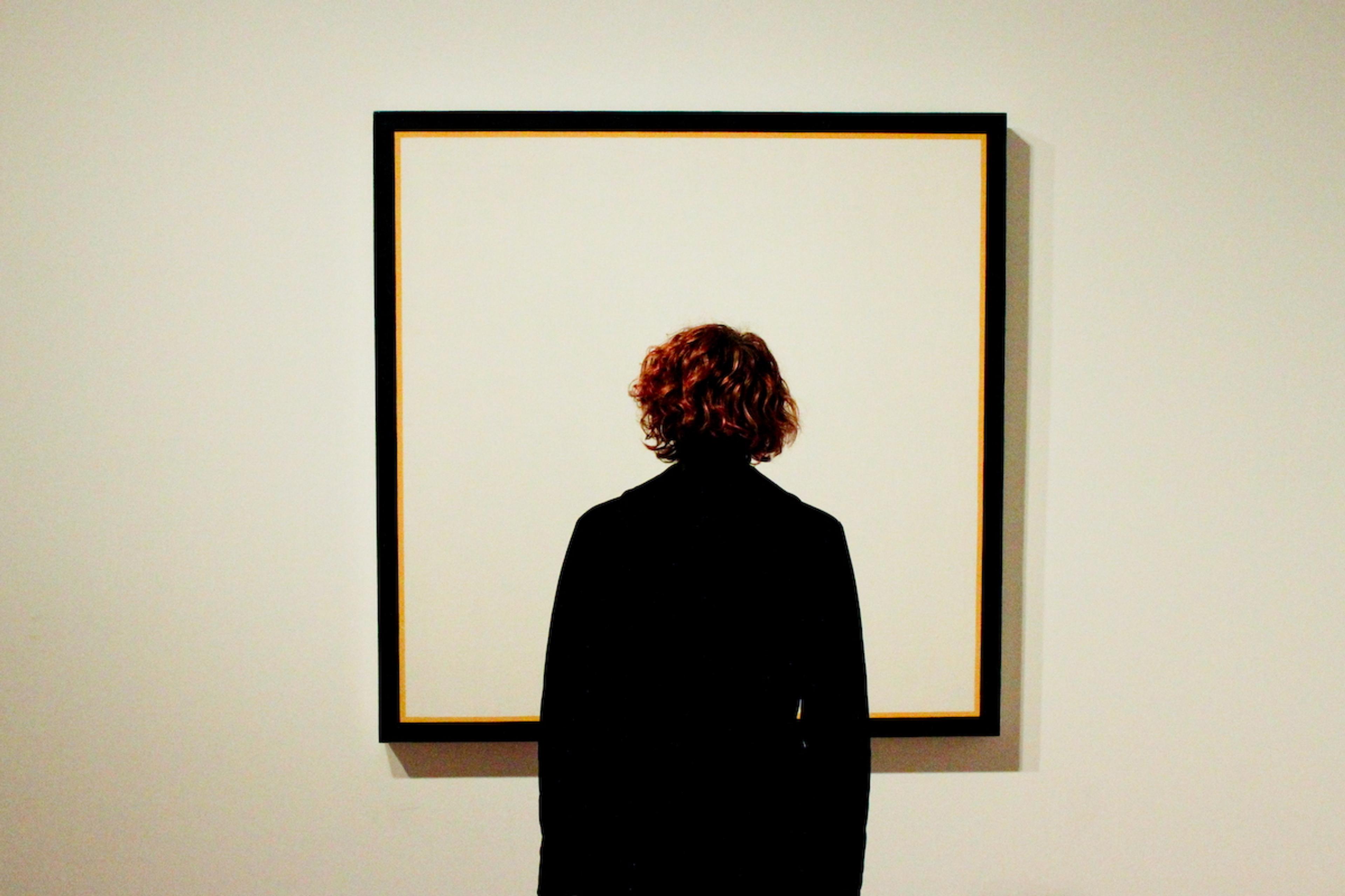 Photo of a person with curly hair viewing a large, square, framed blank canvas on a white wall in a gallery.