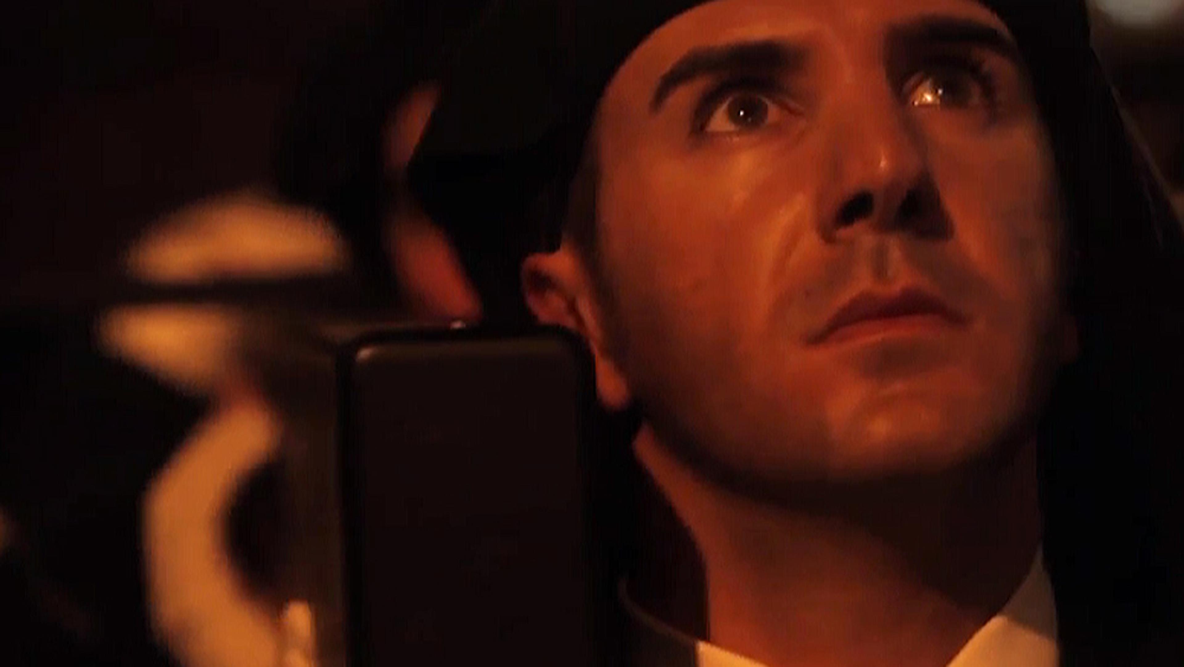 Close-up of a person wearing a dark headscarf looking upwards with a serious expression, lit by warm light.