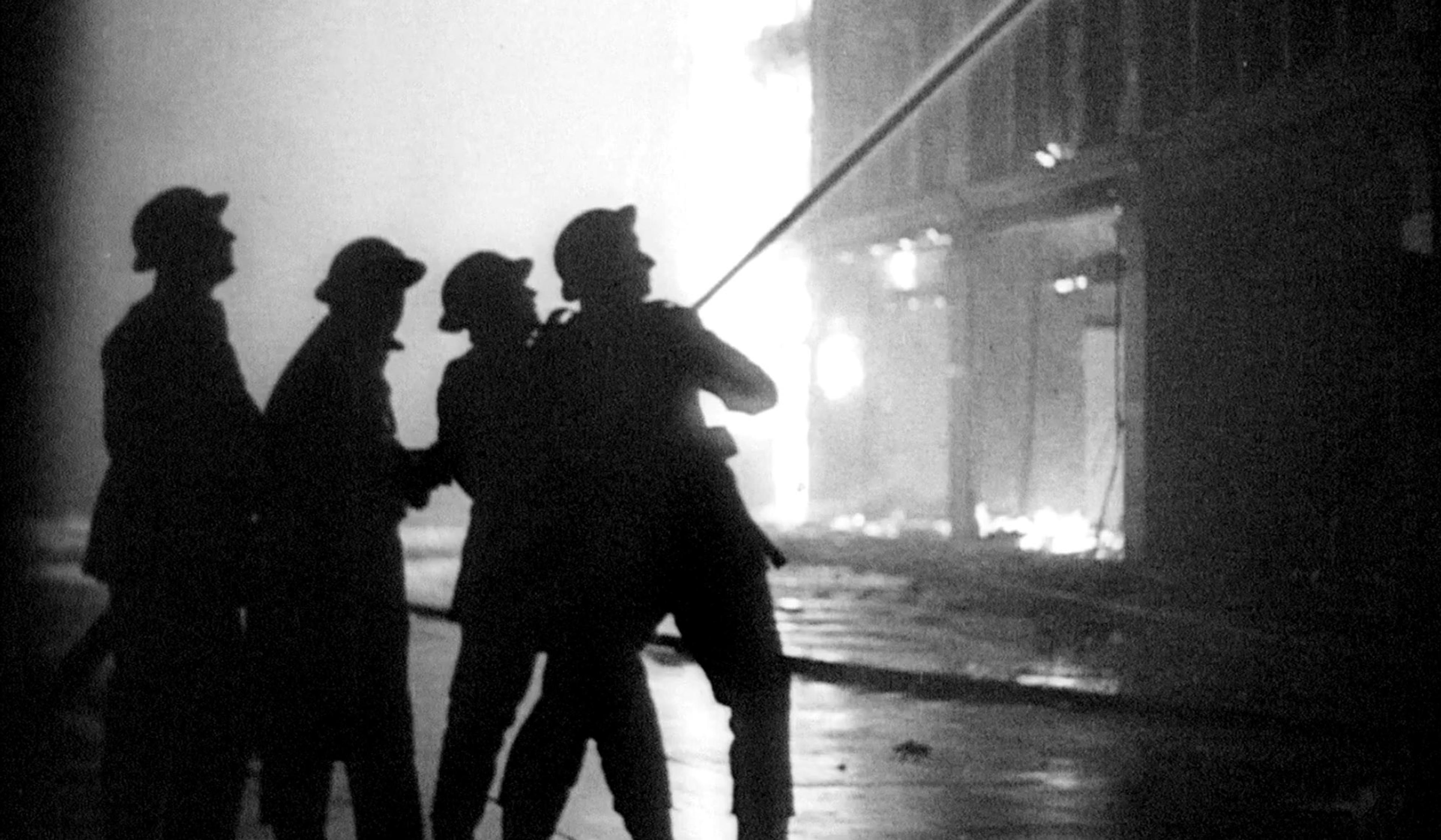 Black-and-white photo of firefighters in silhouette spraying water at a burning building at night, with flames and smoke in the background.